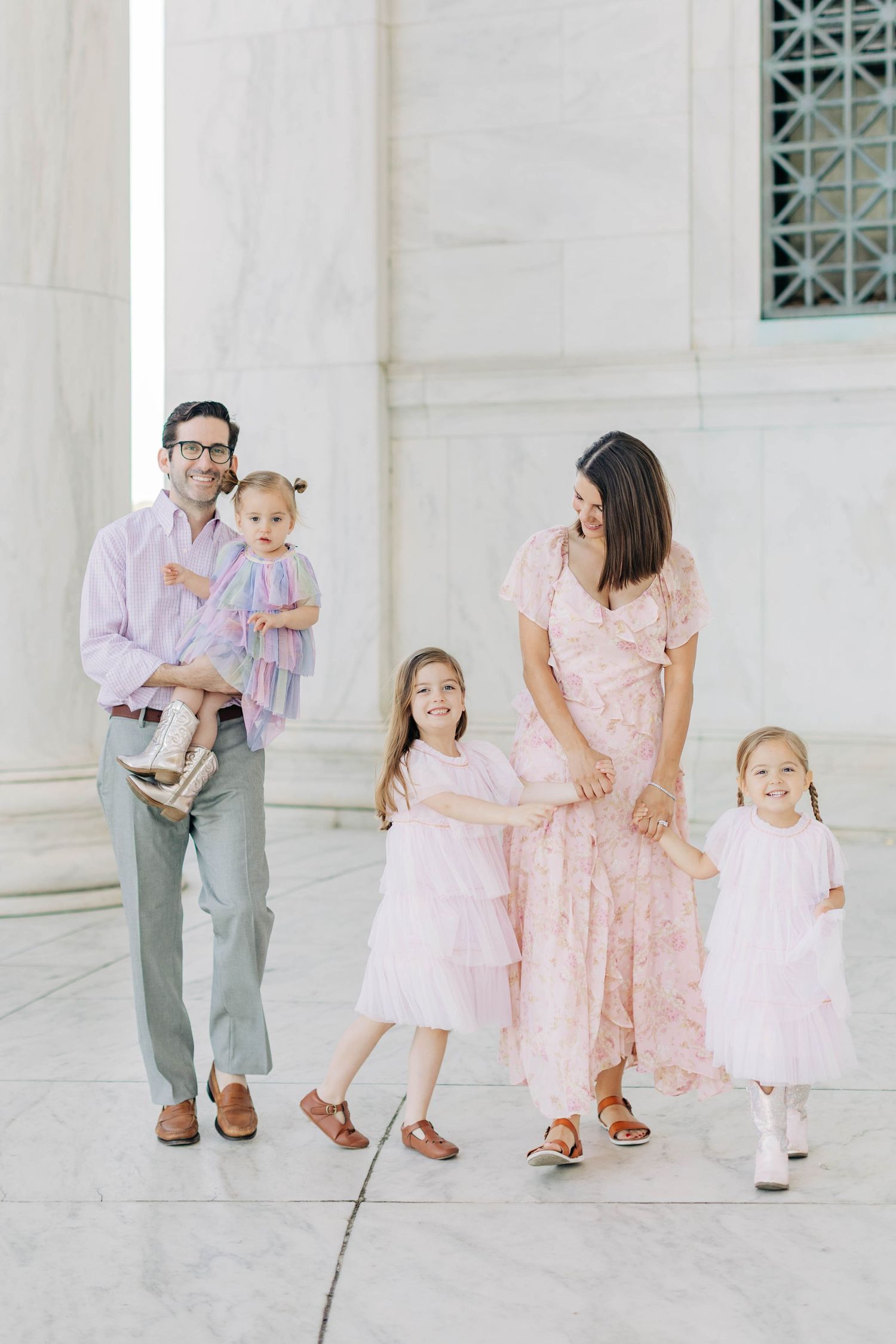A happy family in coordinating pink outfits poses outdoors near marble columns, with dad holding a baby and mom with two girls.