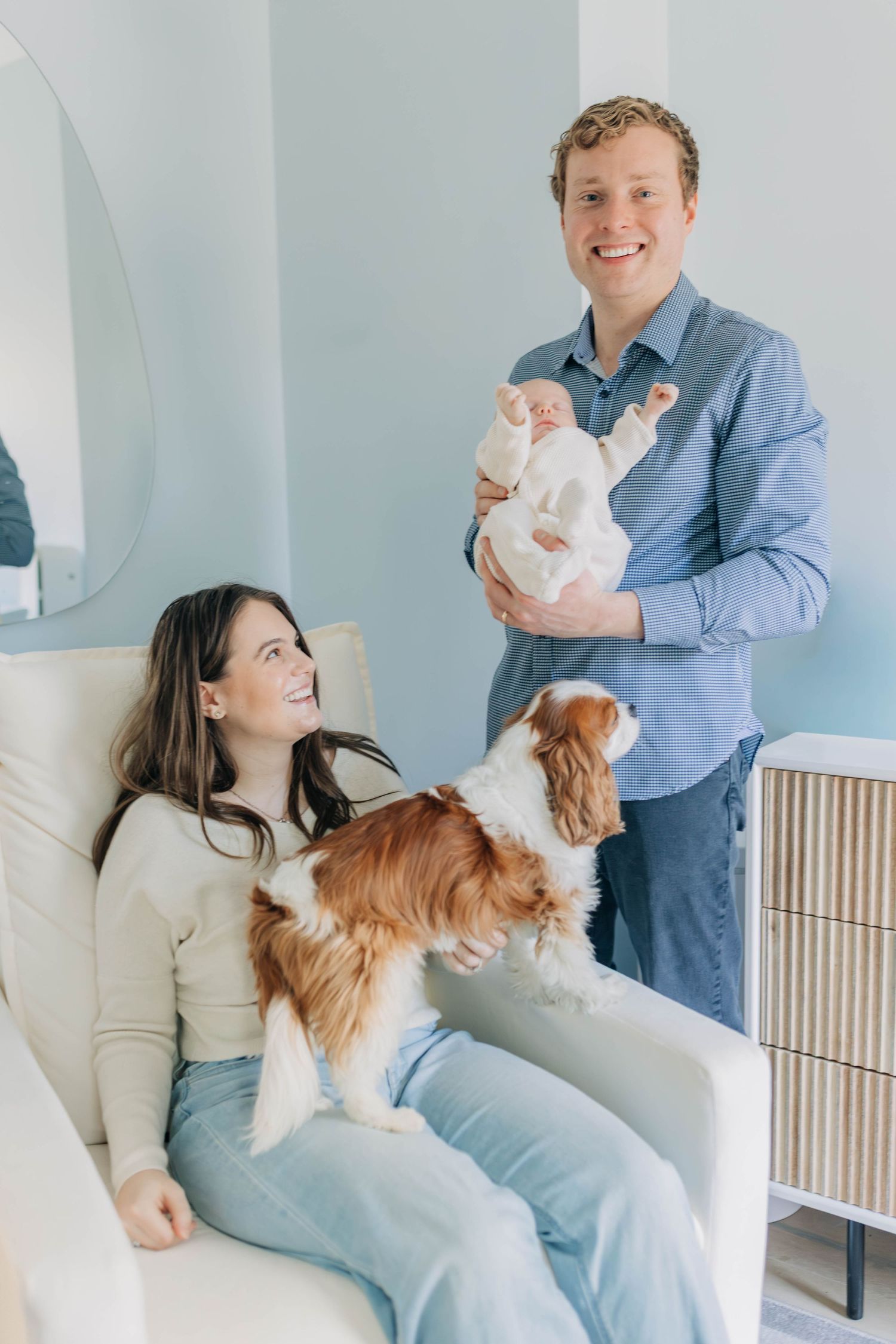 Father holding newborn while mother sits with Cavalier King Charles Spaniel in nursery.