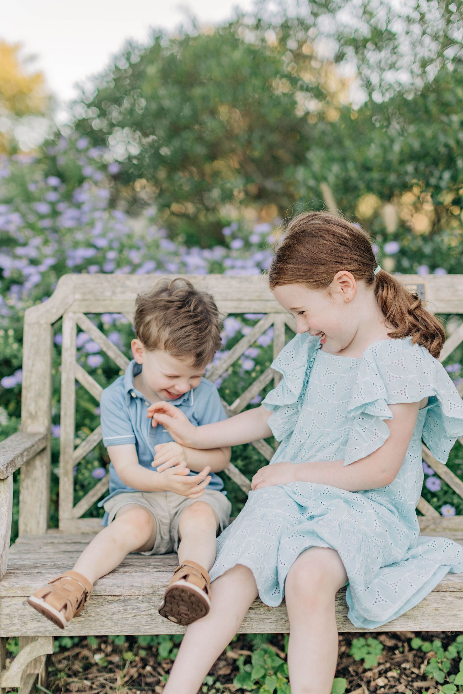 Two young siblings sit on a wooden garden bench surrounded by purple flowers, sharing a joyful moment together.