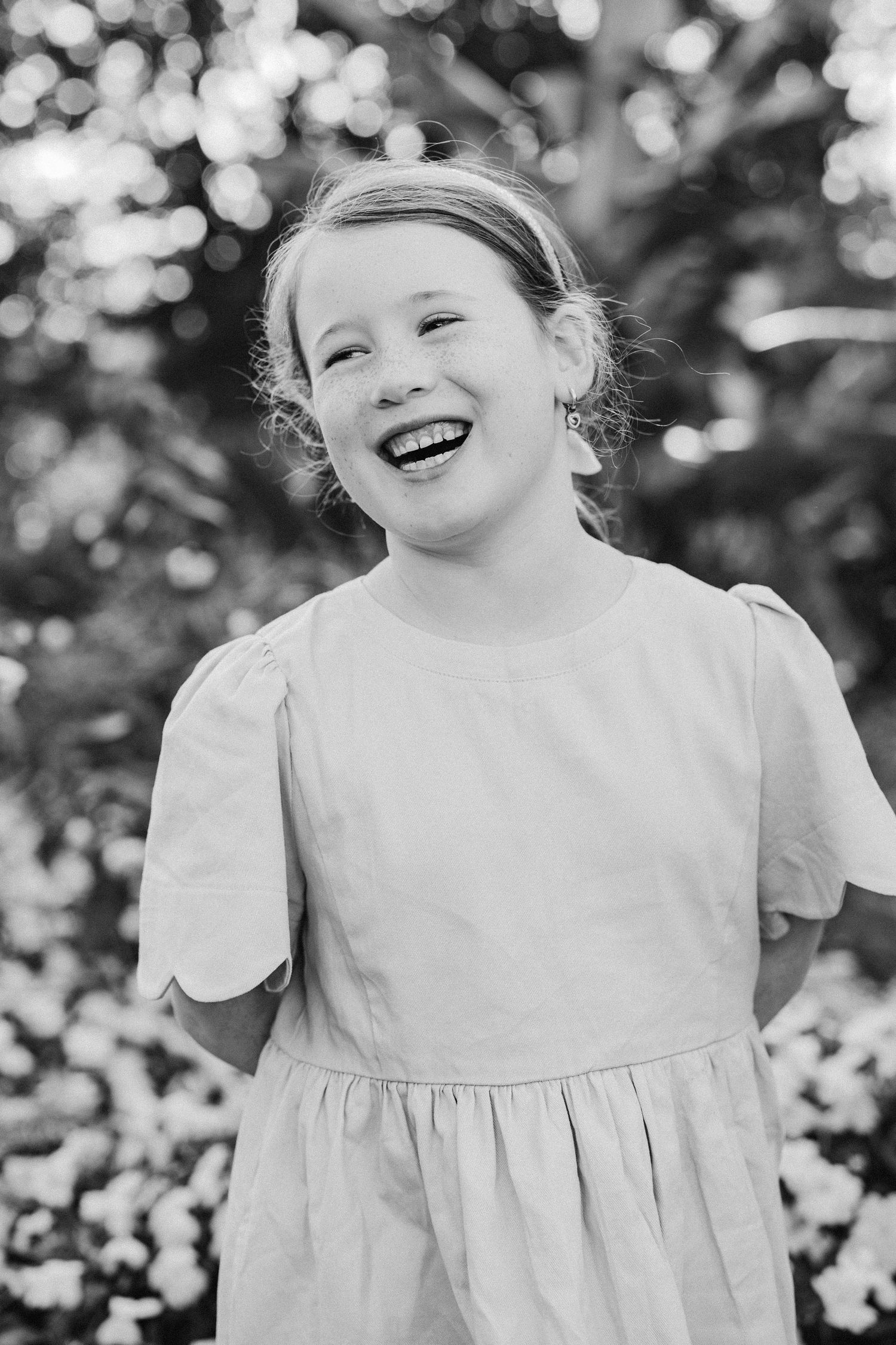 Black and white portrait of a laughing young girl in a dress, standing outdoors among flowers.