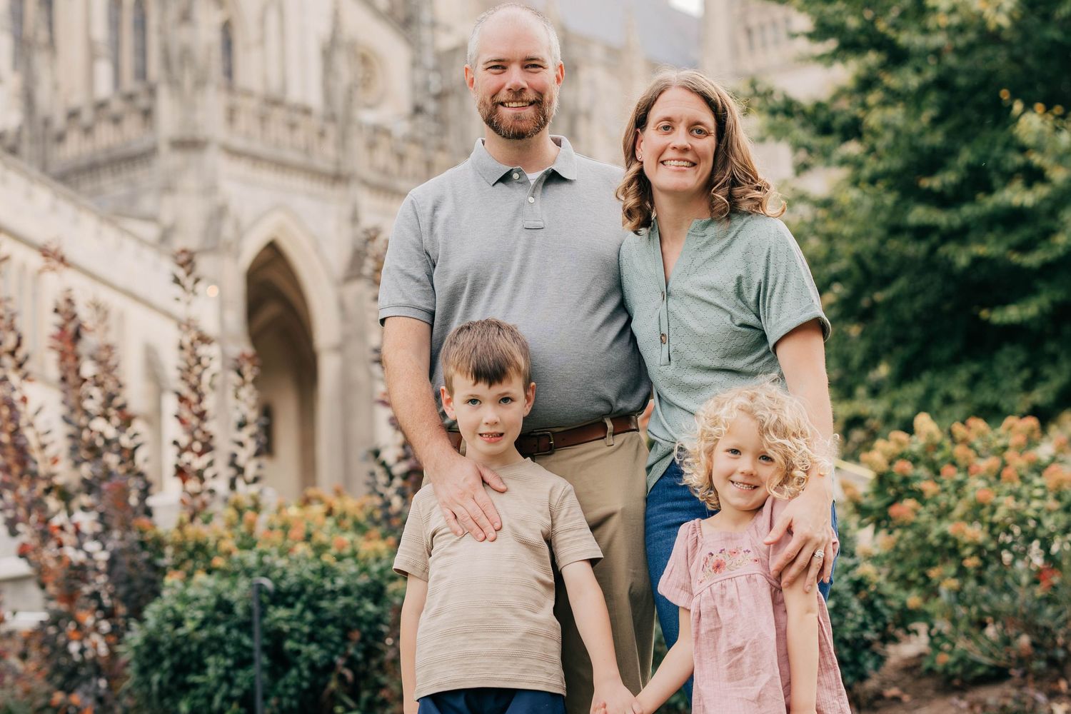 Smiling family of four in coordinated neutral outfits posing outdoors in front of a historic Gothic stone building surrounded by greenery.