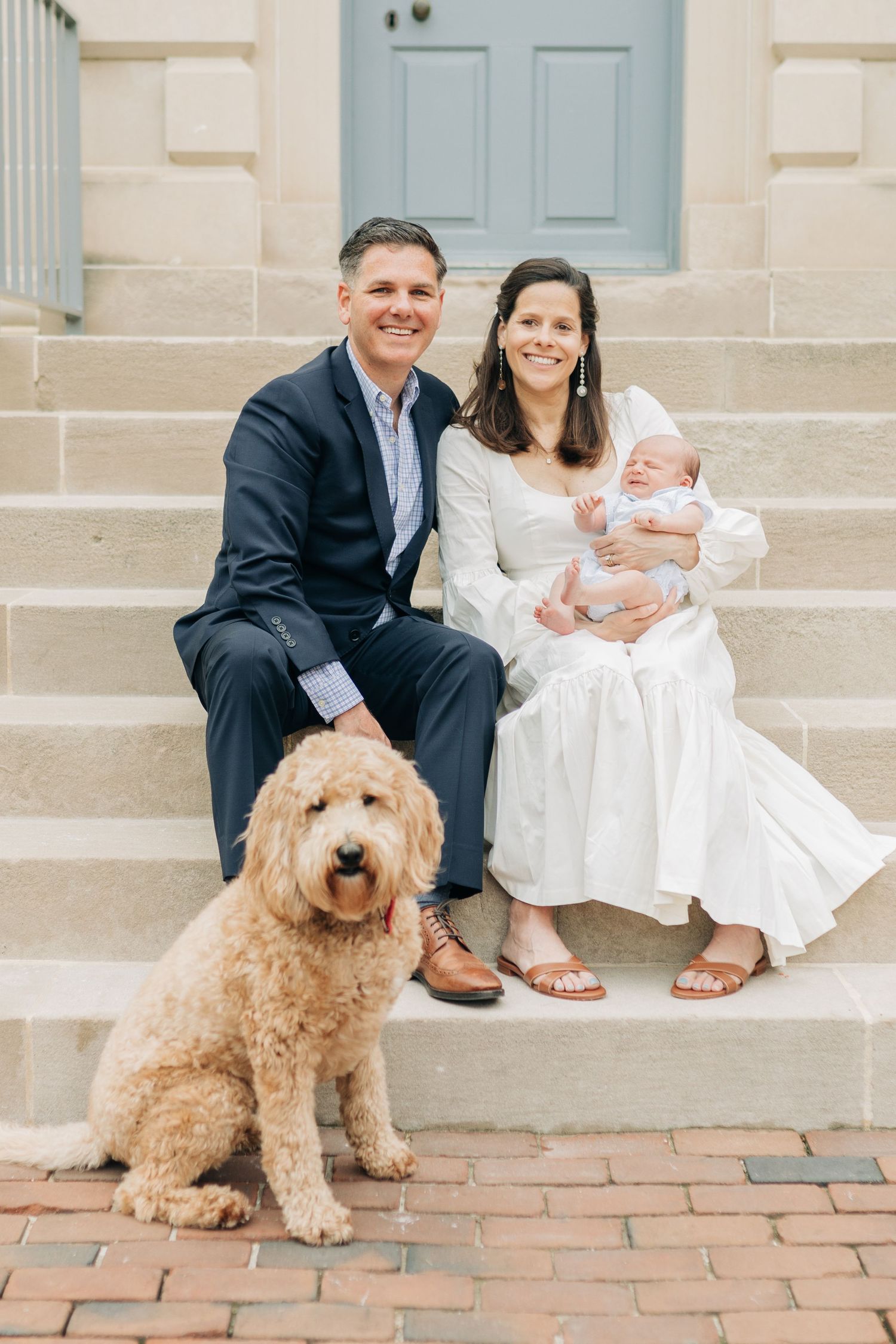A family sits on stone steps with their goldendoodle dog in front of a building with blue doors.