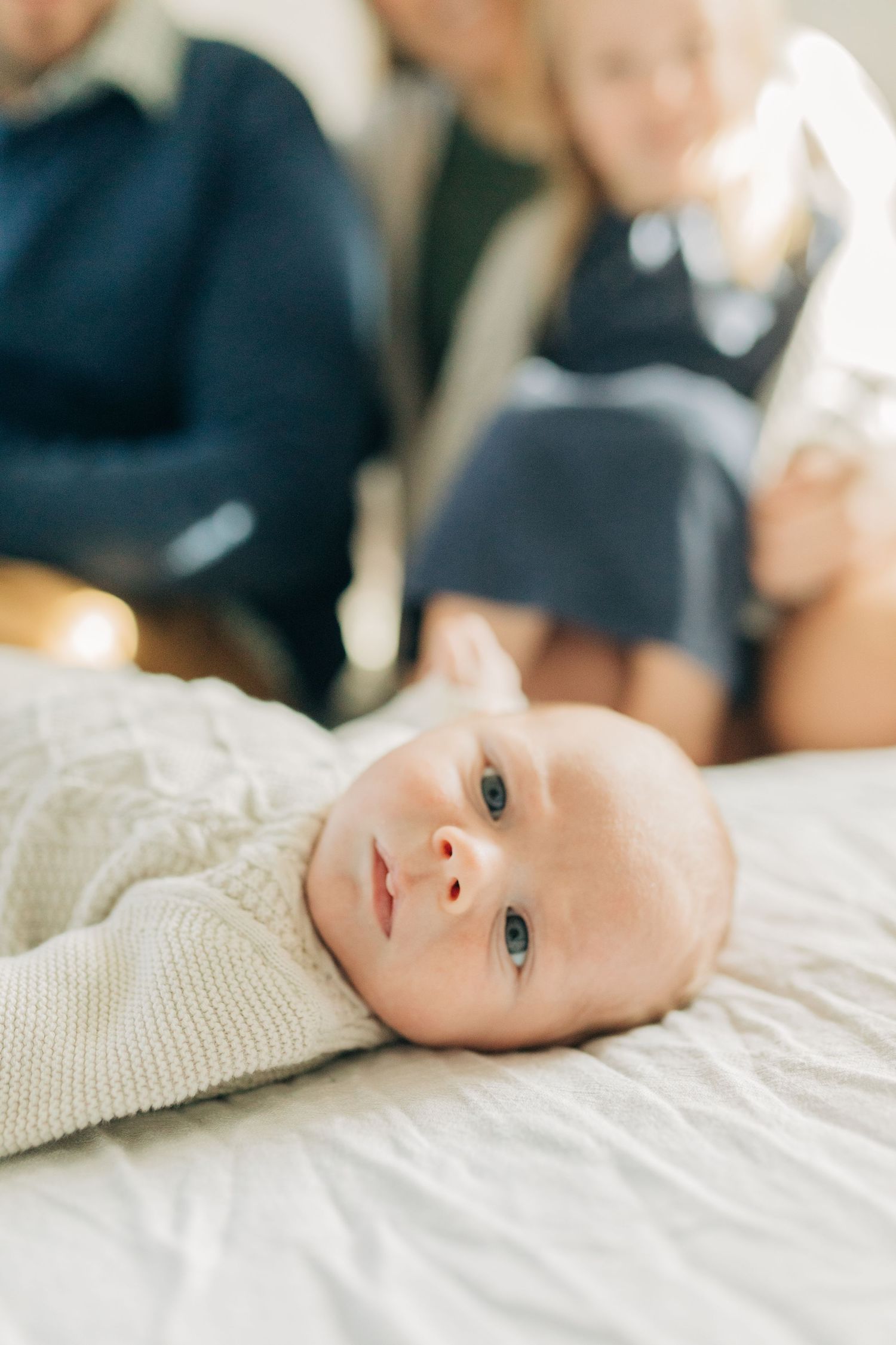 A baby in cream sweater lies on white bedding looking at camera.