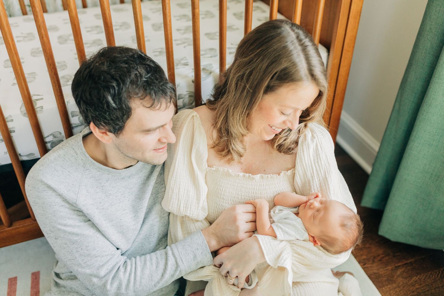 Parents share an intimate moment with their newborn baby in a nursery with green curtains and wooden crib.