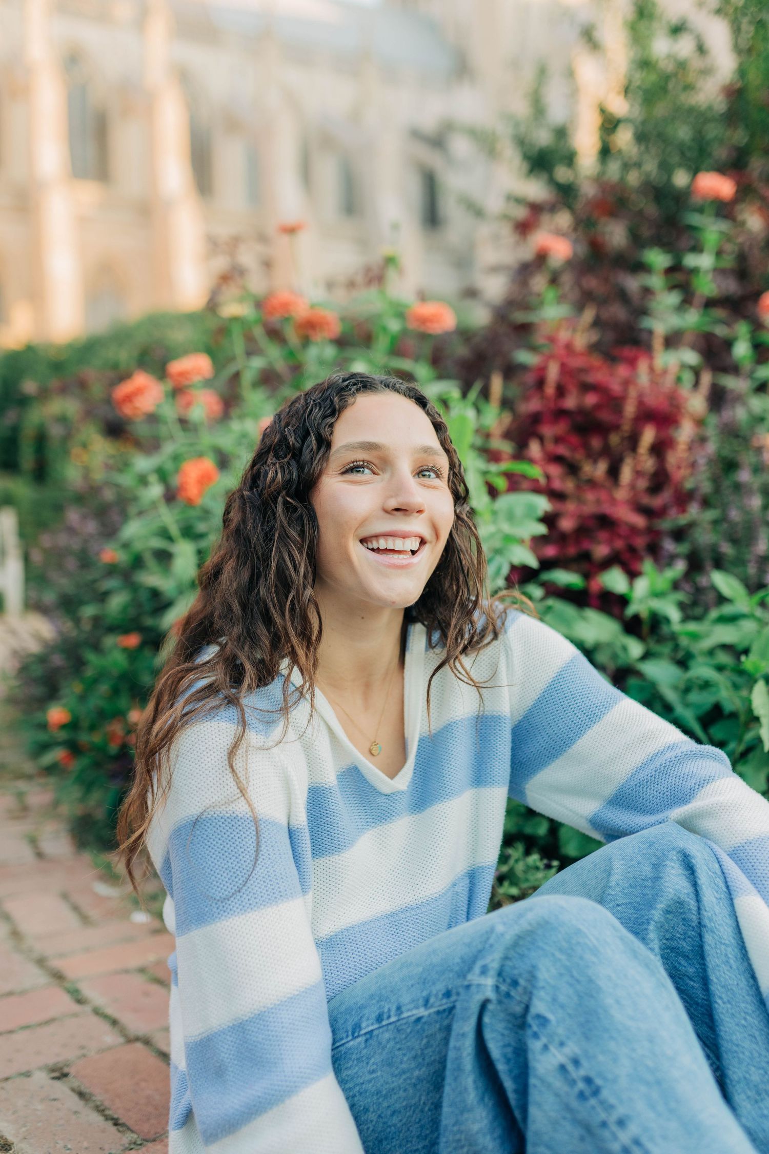 A person in casual wear smiles while sitting among colorful garden flowers and foliage.