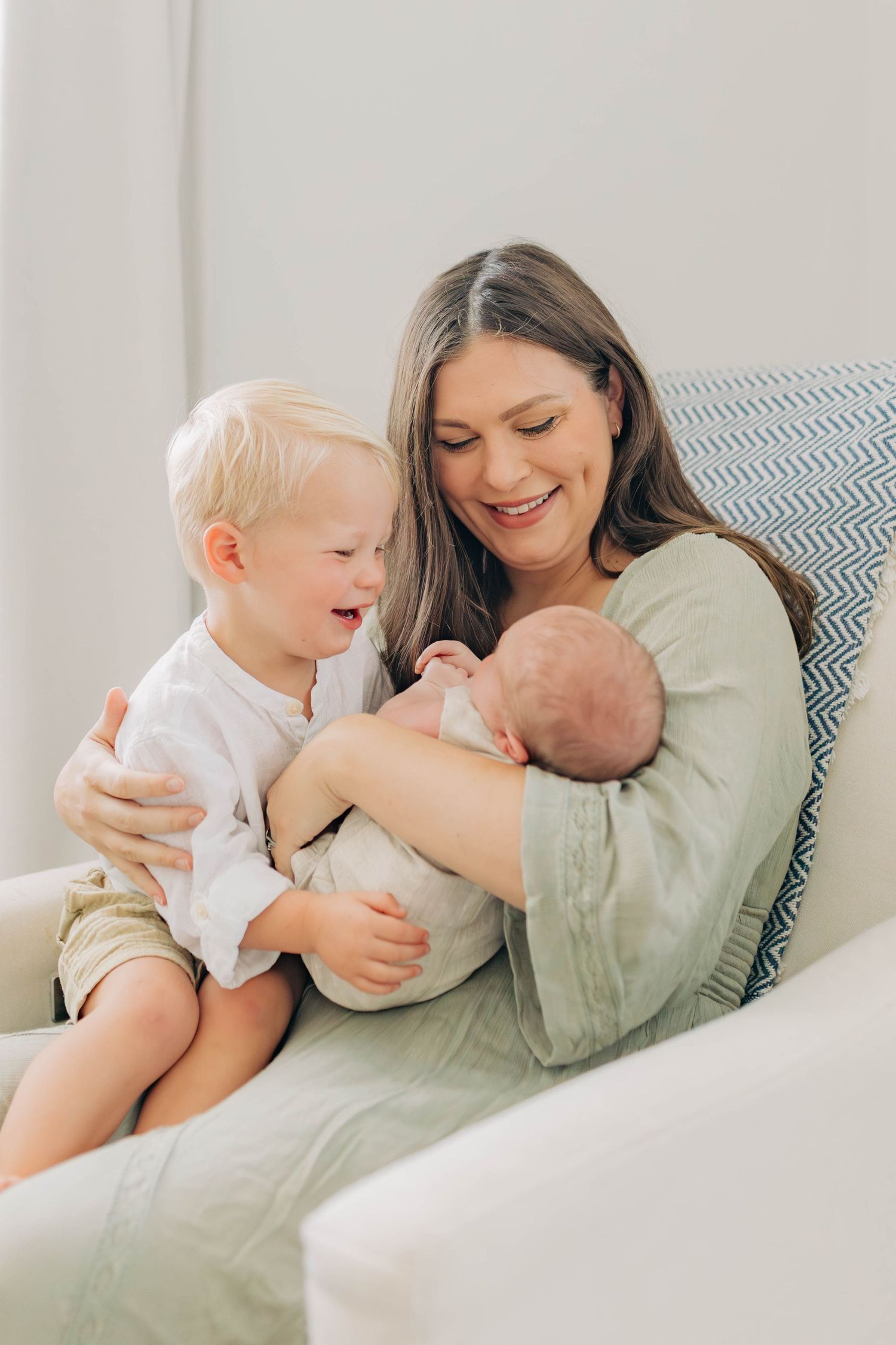 A mother cuddles with her baby and toddler on a cozy couch.