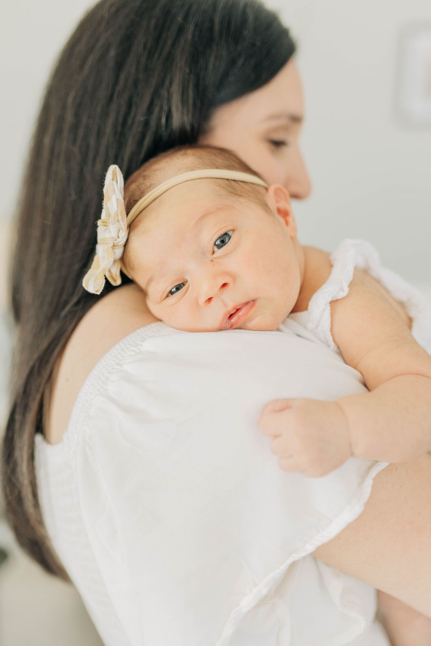Baby in white outfit with bow headband resting against caregiver's shoulder.