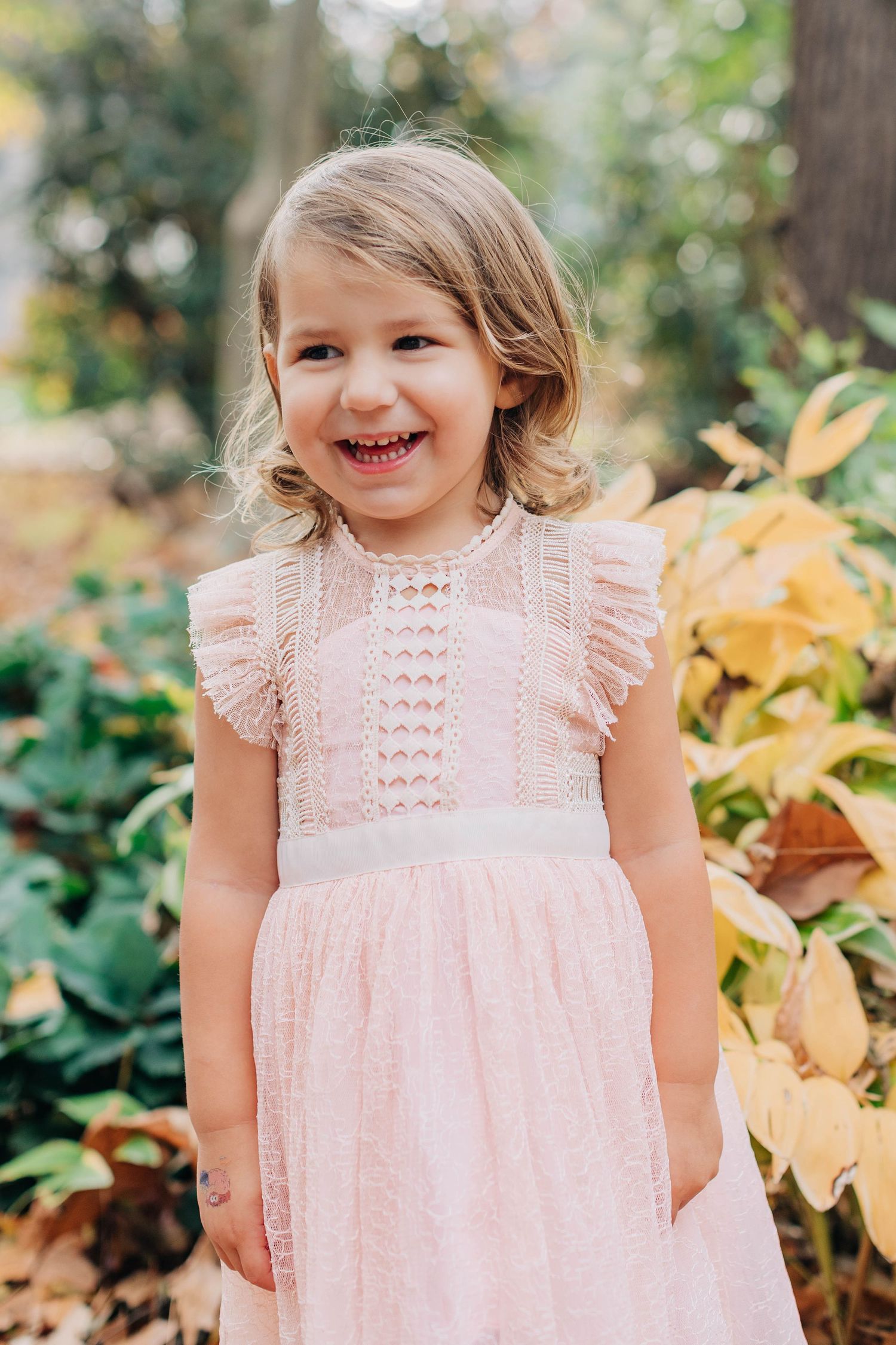 A child wearing a pink eyelet lace dress smiles joyfully in an outdoor garden setting.