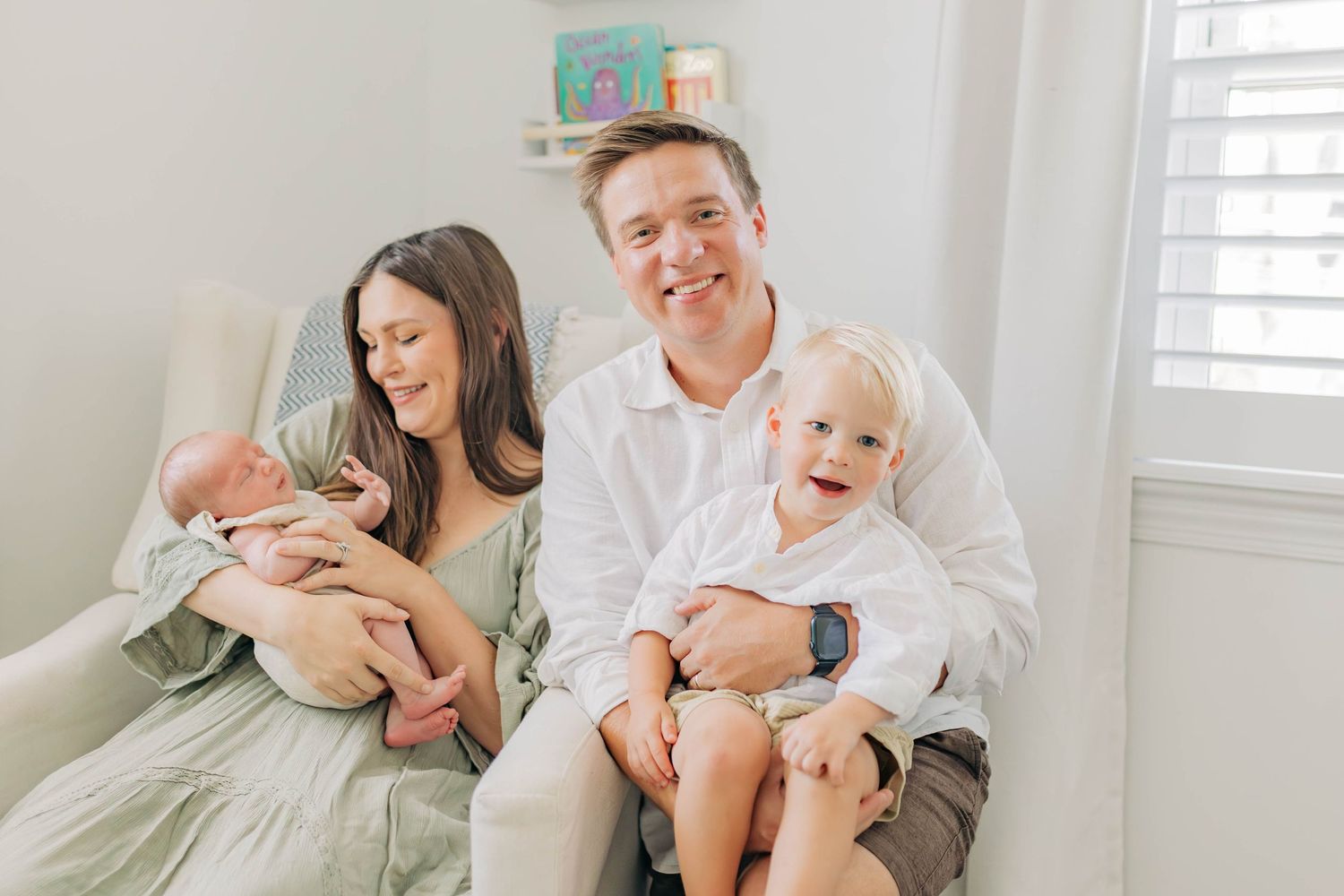 A family lounges together on a bed in a bright room with white decor and window shutters.