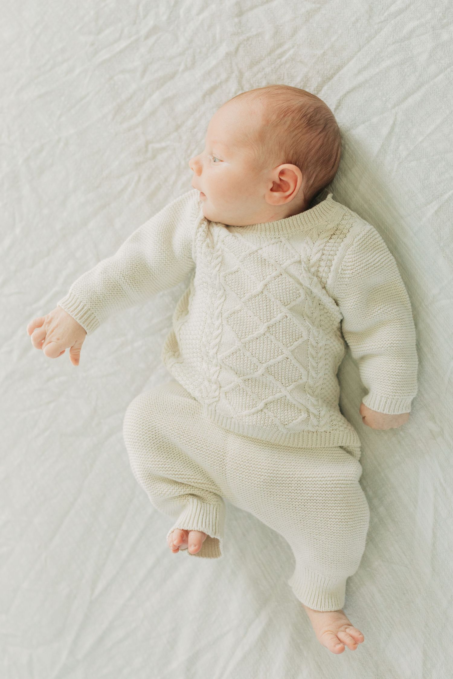 Infant wearing a cream knit sweater lies on white textured bedding.
