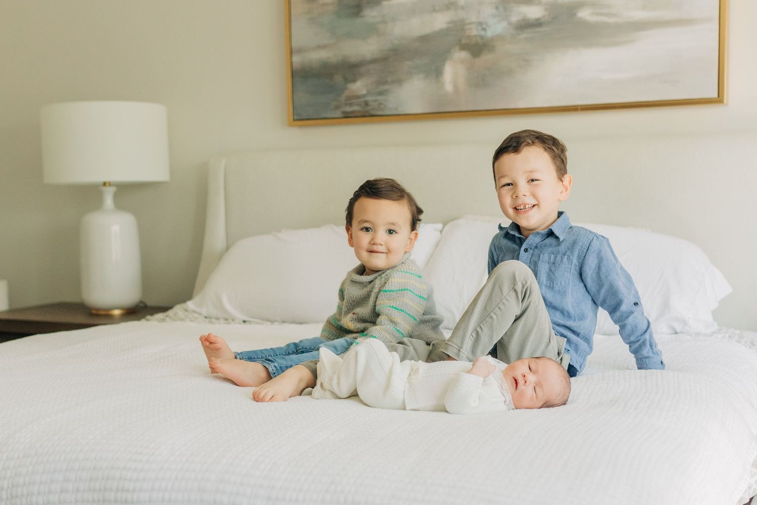 Siblings bond together while relaxing on white bedding beneath modern wall art.