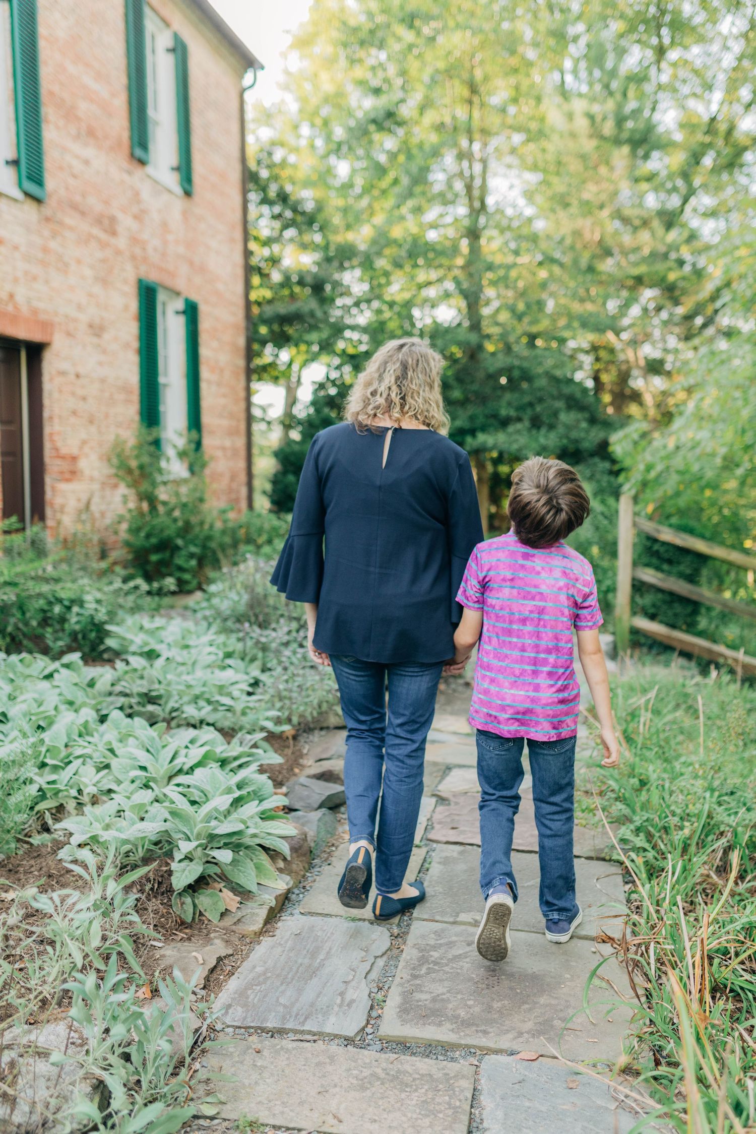Two people walking away on stone path beside lush garden and brick building.