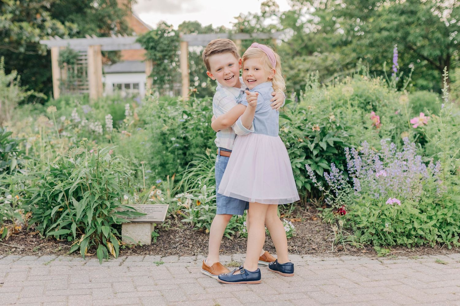 Two young children in coordinating blue and pink outfits share playful moments in a garden setting.