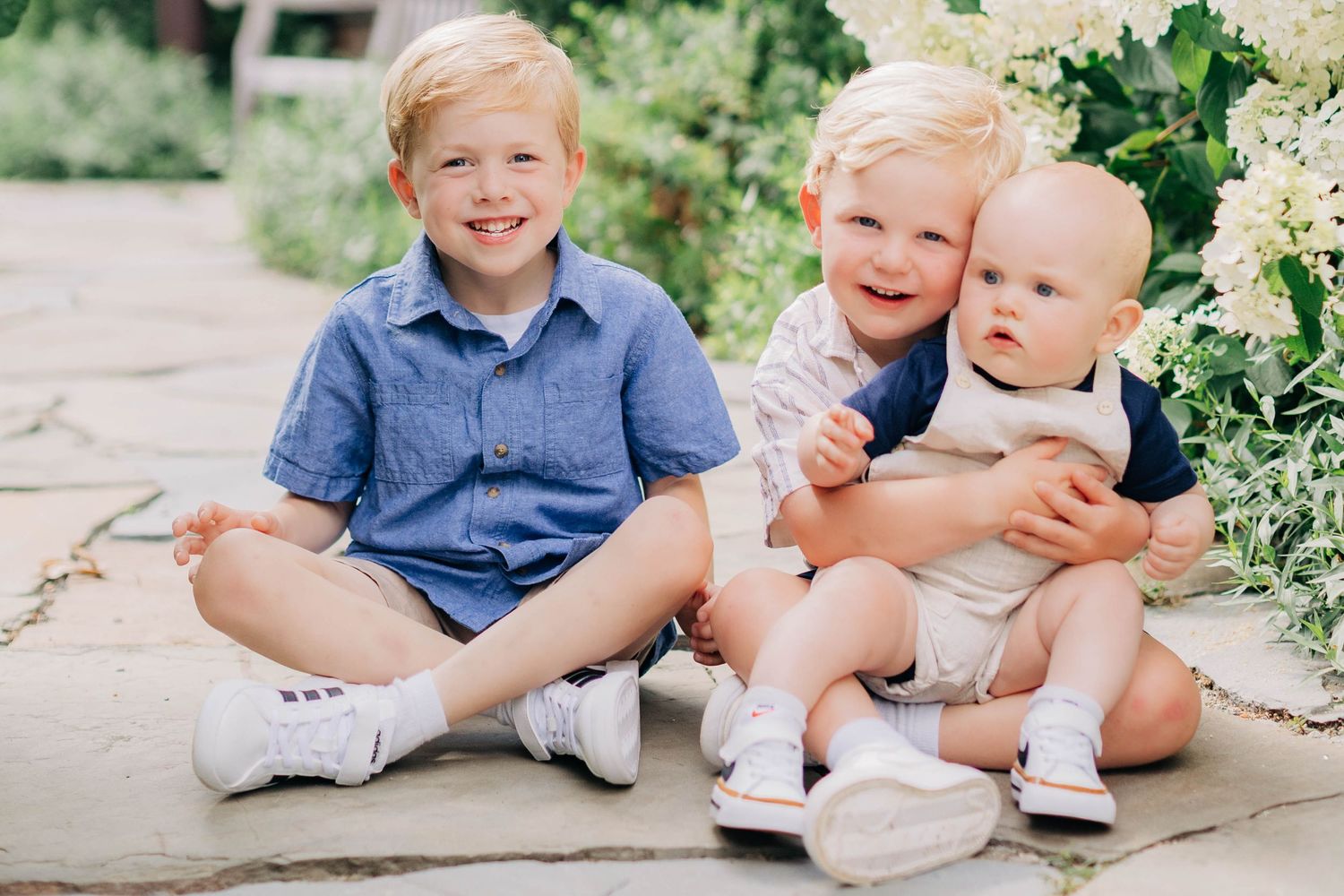 Three young blonde brothers sitting together outdoors, smiling, with white hydrangeas in the background.