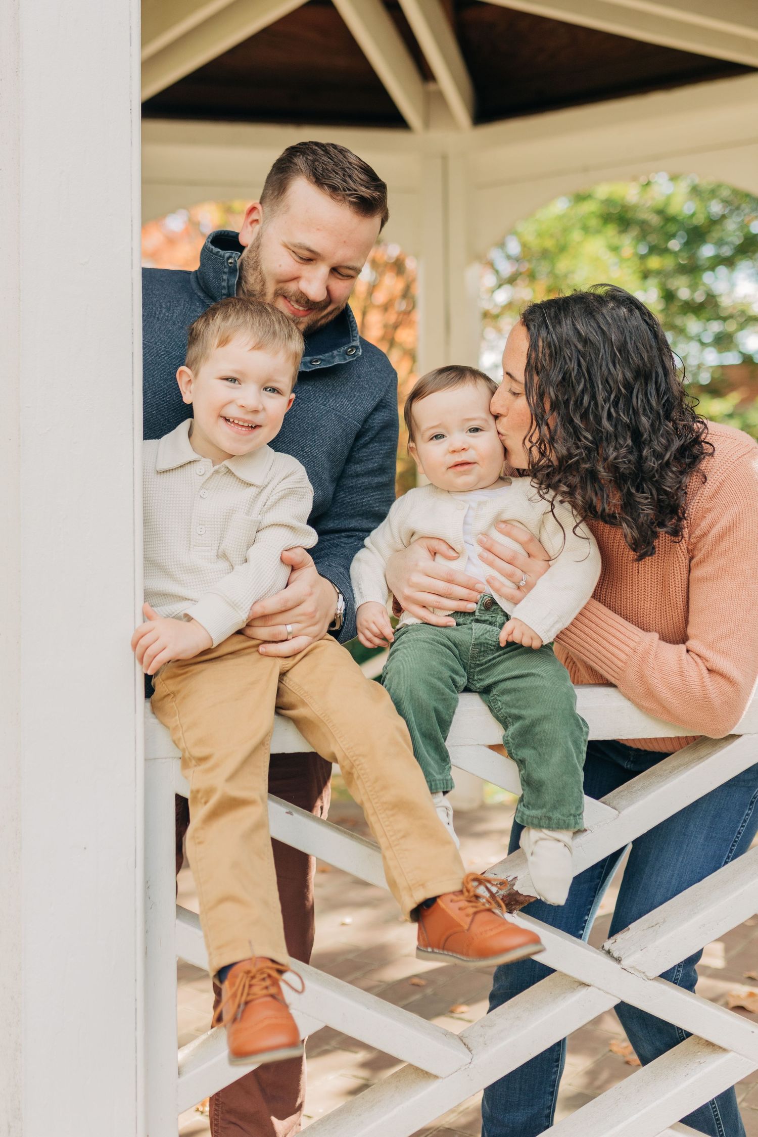 Happy family of four posing in a gazebo, parents smiling at two young boys in tan and green pants.