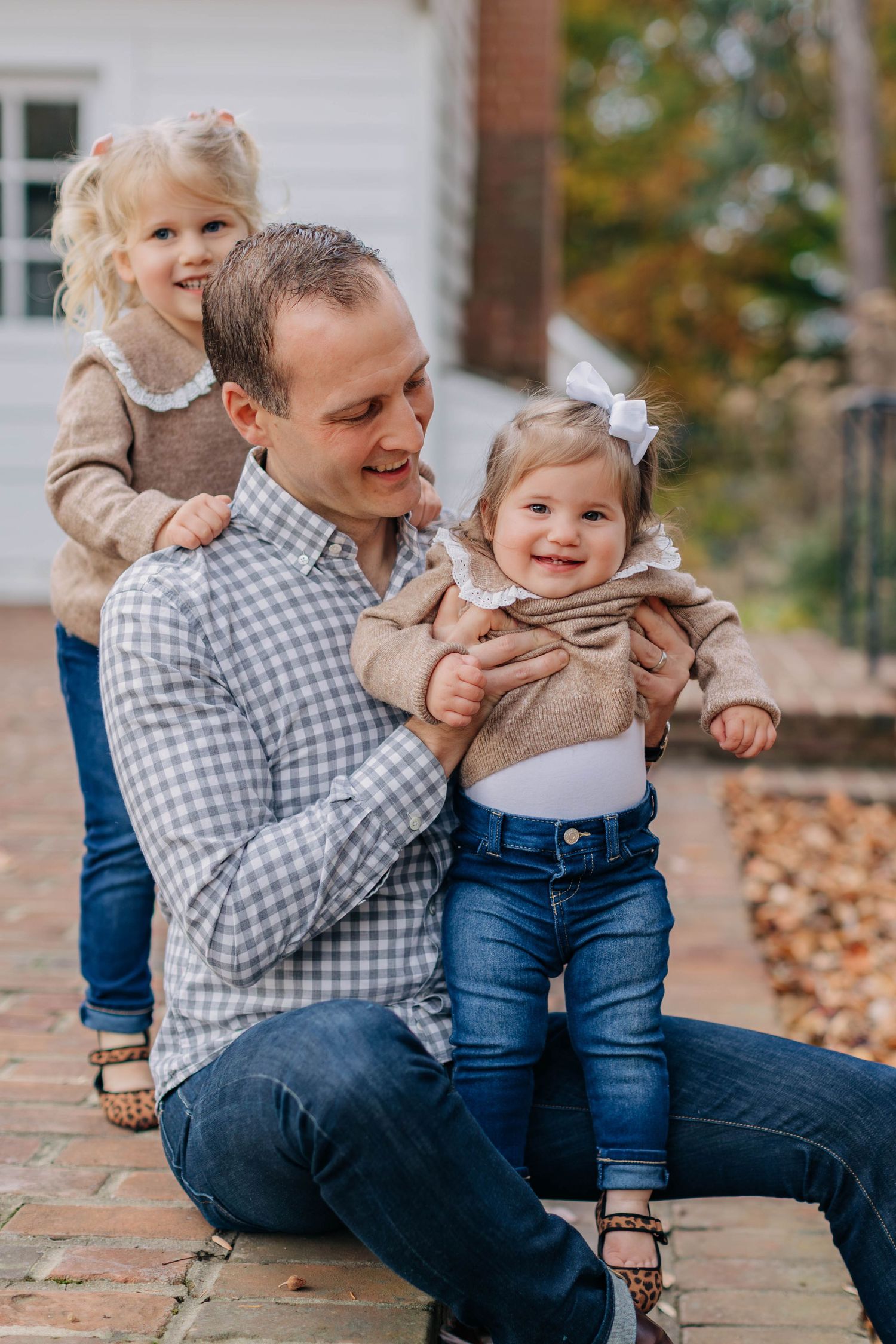 A father smiles while holding two young blonde girls in jeans and tan sweaters outdoors on a brick patio.