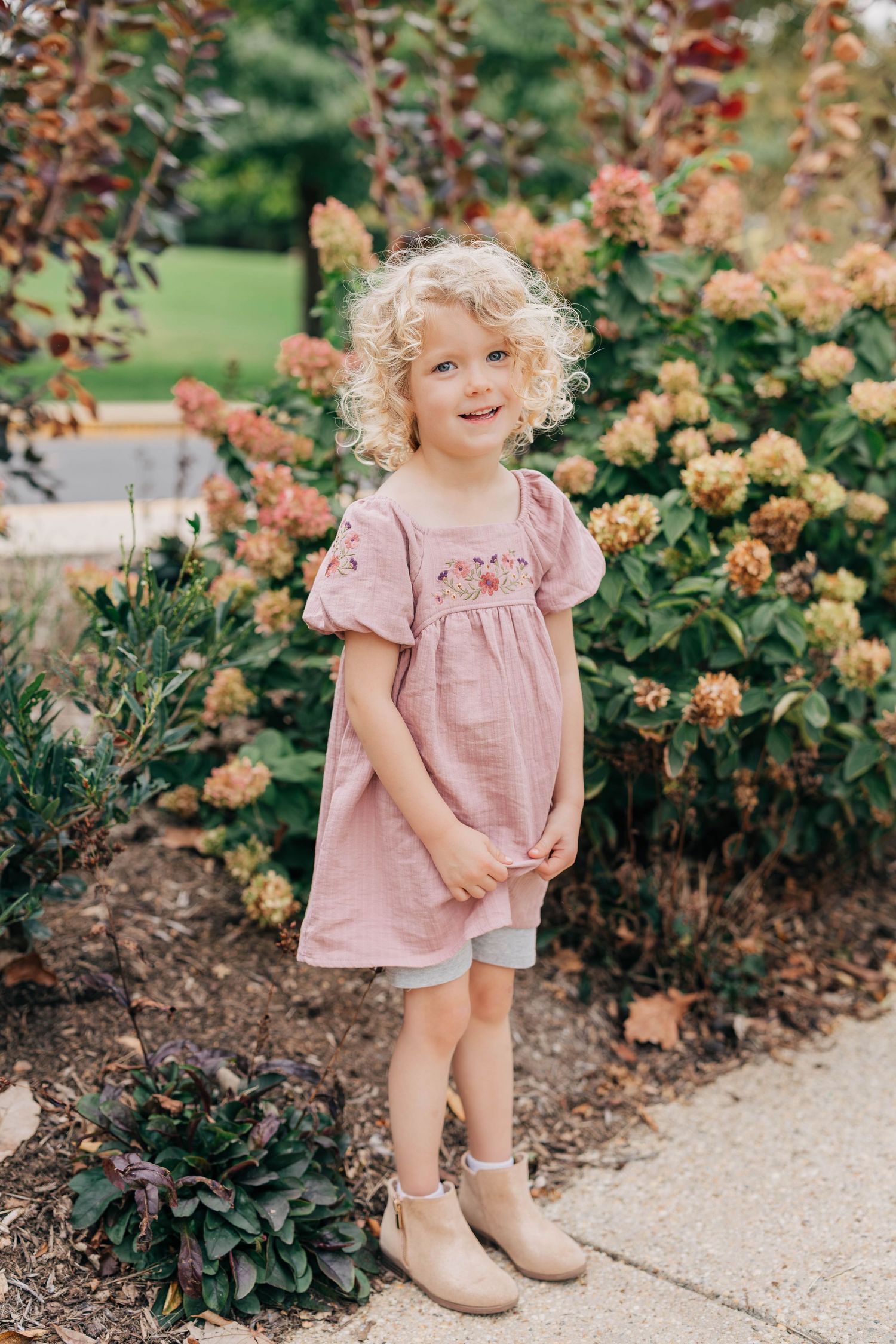 Smiling toddler with curly blonde hair in a pink linen dress and tan ankle boots standing by blooming hydrangeas in a garden.