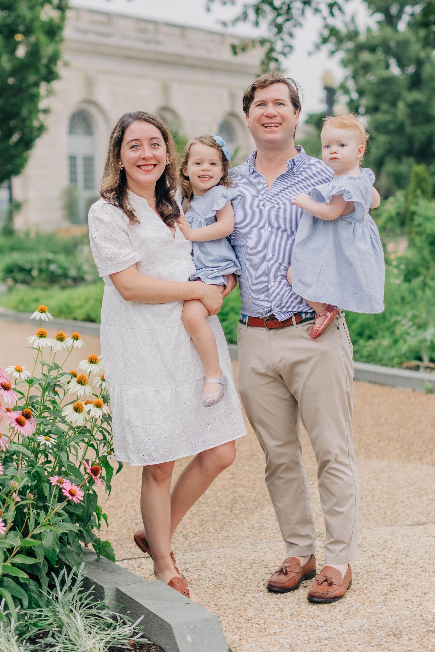A smiling family of four poses outdoors near a garden, parents holding two young girls in matching blue dresses.
