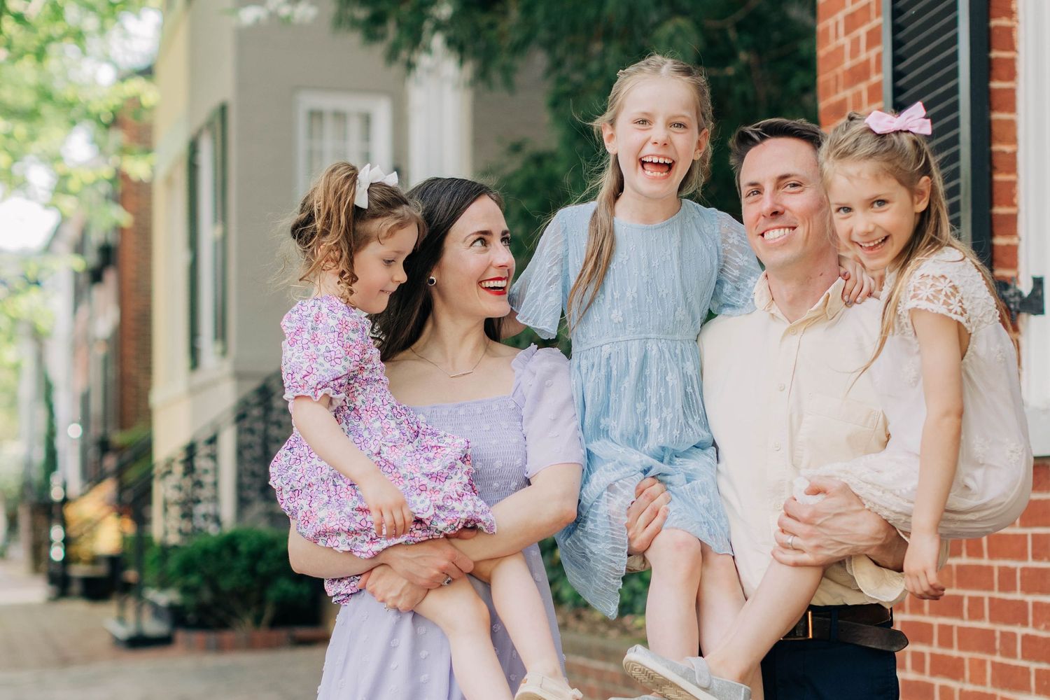 Happy family of five posing outdoors on a brick sidewalk, parents holding three young daughters in pastel dresses.