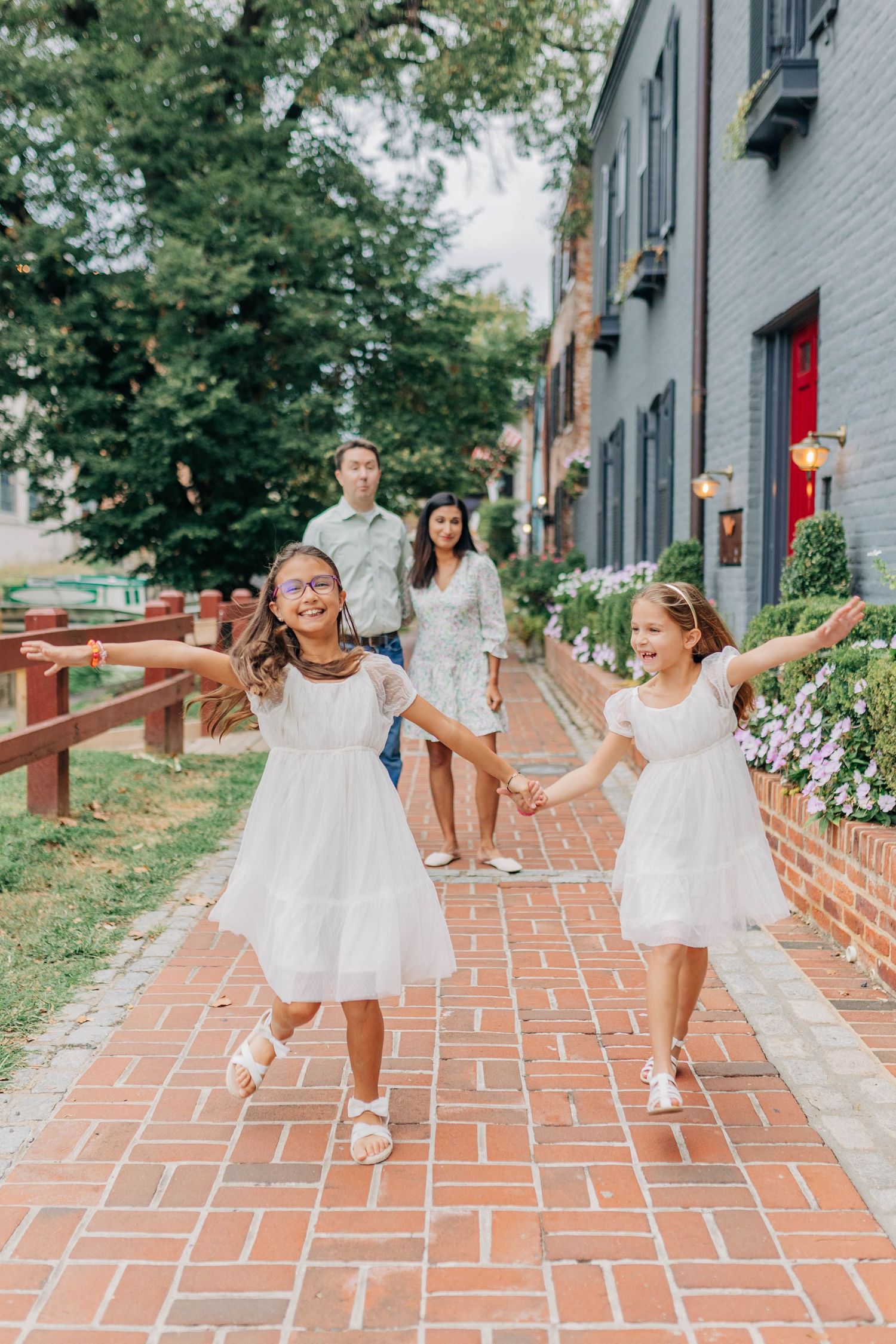 Two girls in white dresses hold hands on a brick path while parents watch in a charming neighborhood setting.