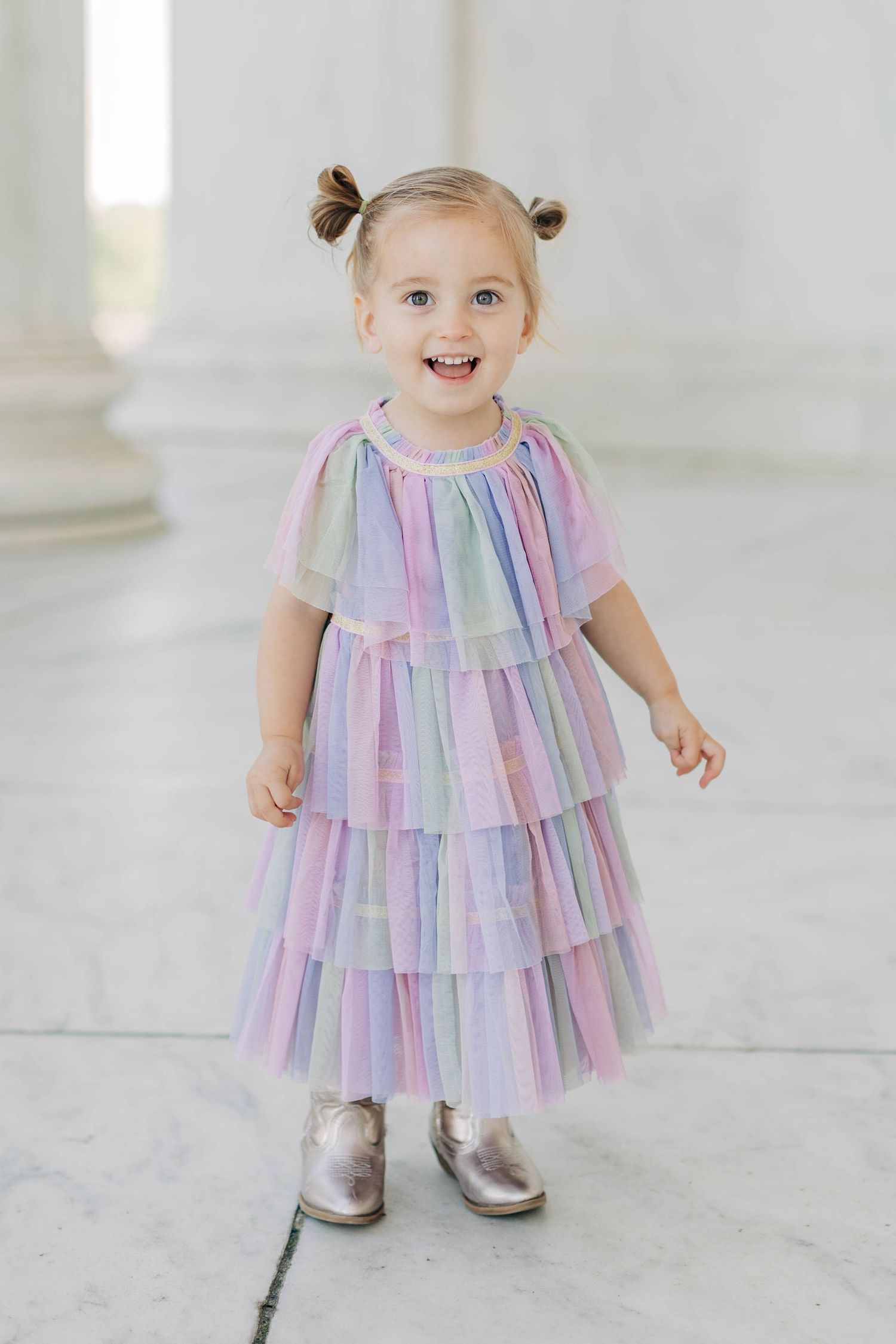 A cheerful toddler girl in a colorful rainbow tiered tulle dress with silver boots smiles brightly.