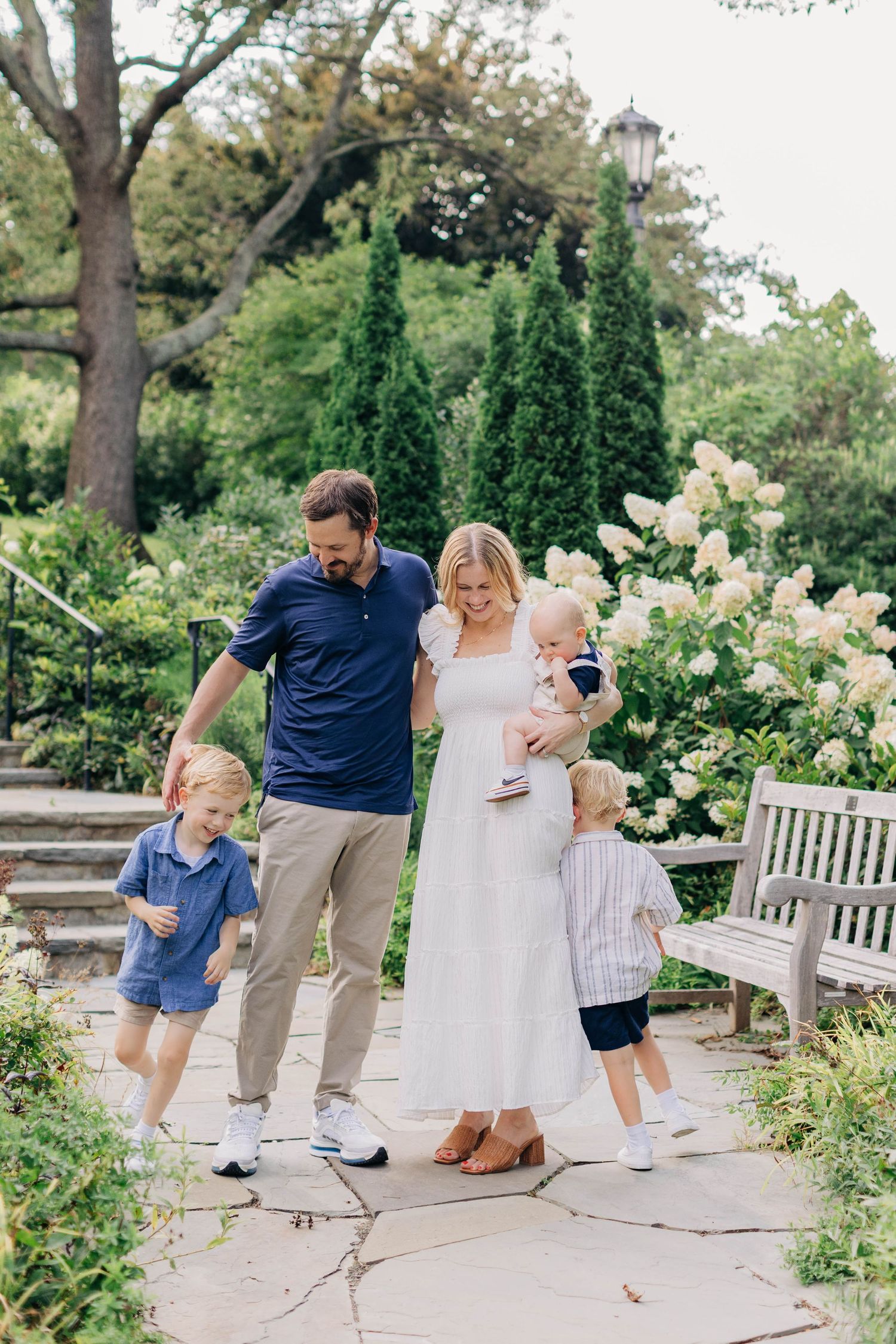 Happy family of four with two young boys walking in a lush garden with white hydrangeas and cypress trees.