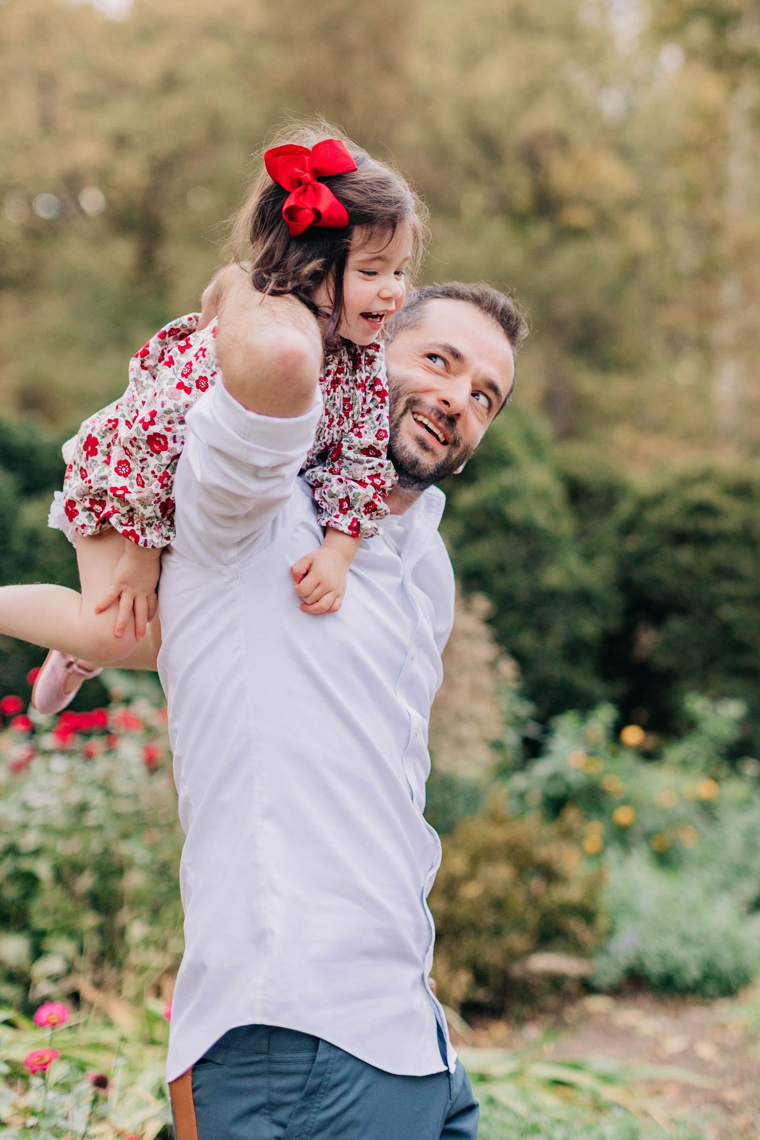 A father joyfully lifts his young daughter in a red floral dress outdoors in a lush garden setting.
