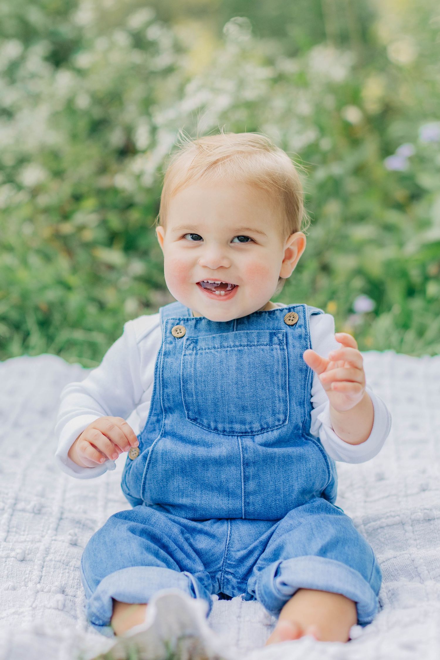 Smiling baby in denim overalls sitting on a blanket outdoors surrounded by green plants and wildflowers.