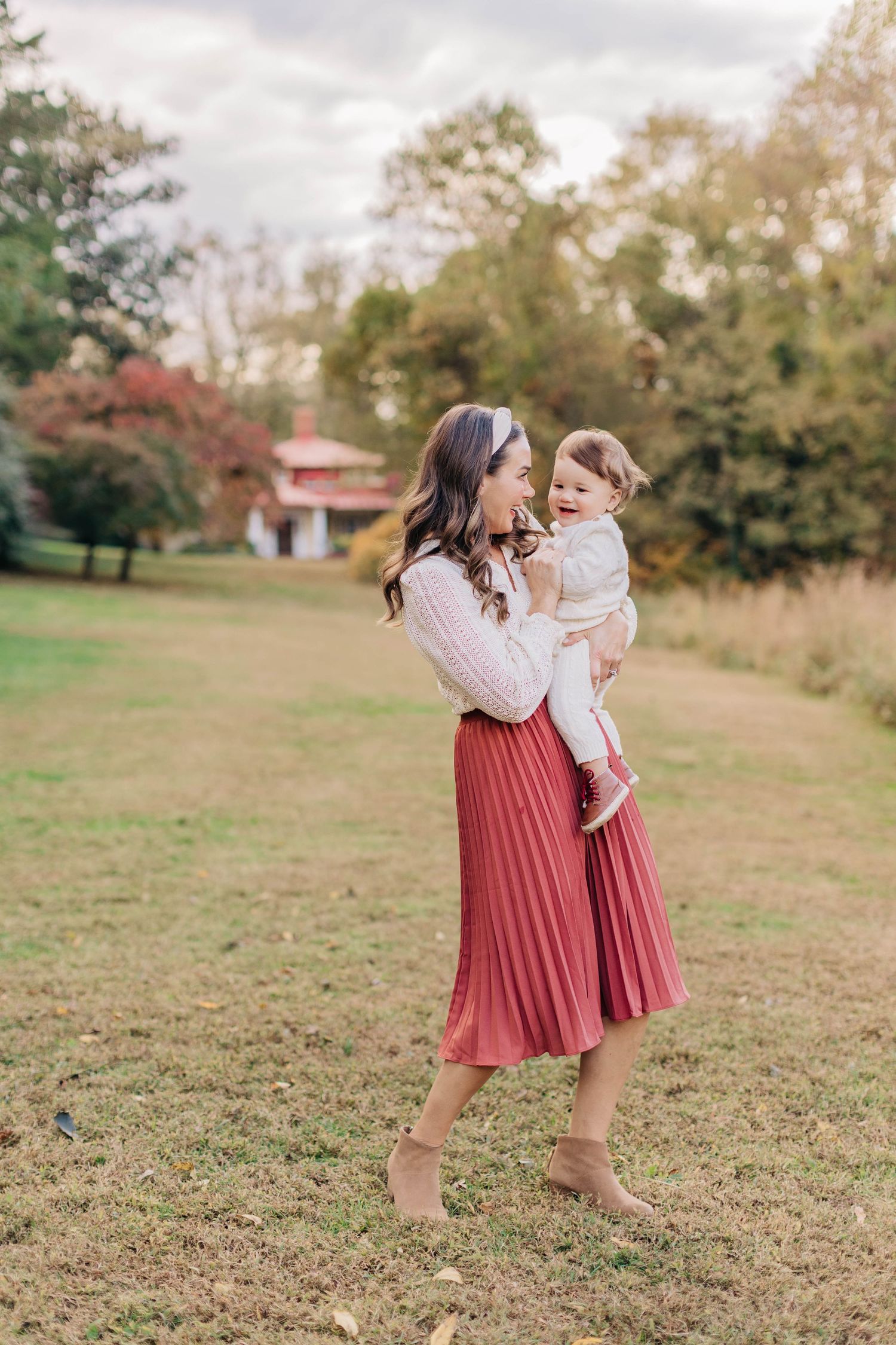 A mother in a red pleated skirt holds her toddler in a lush green park during autumn.