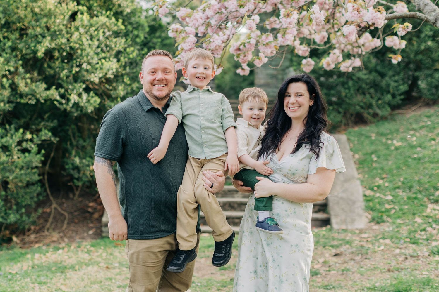 Happy family of four posing outdoors under blooming cherry blossom tree on green lawn in spring.