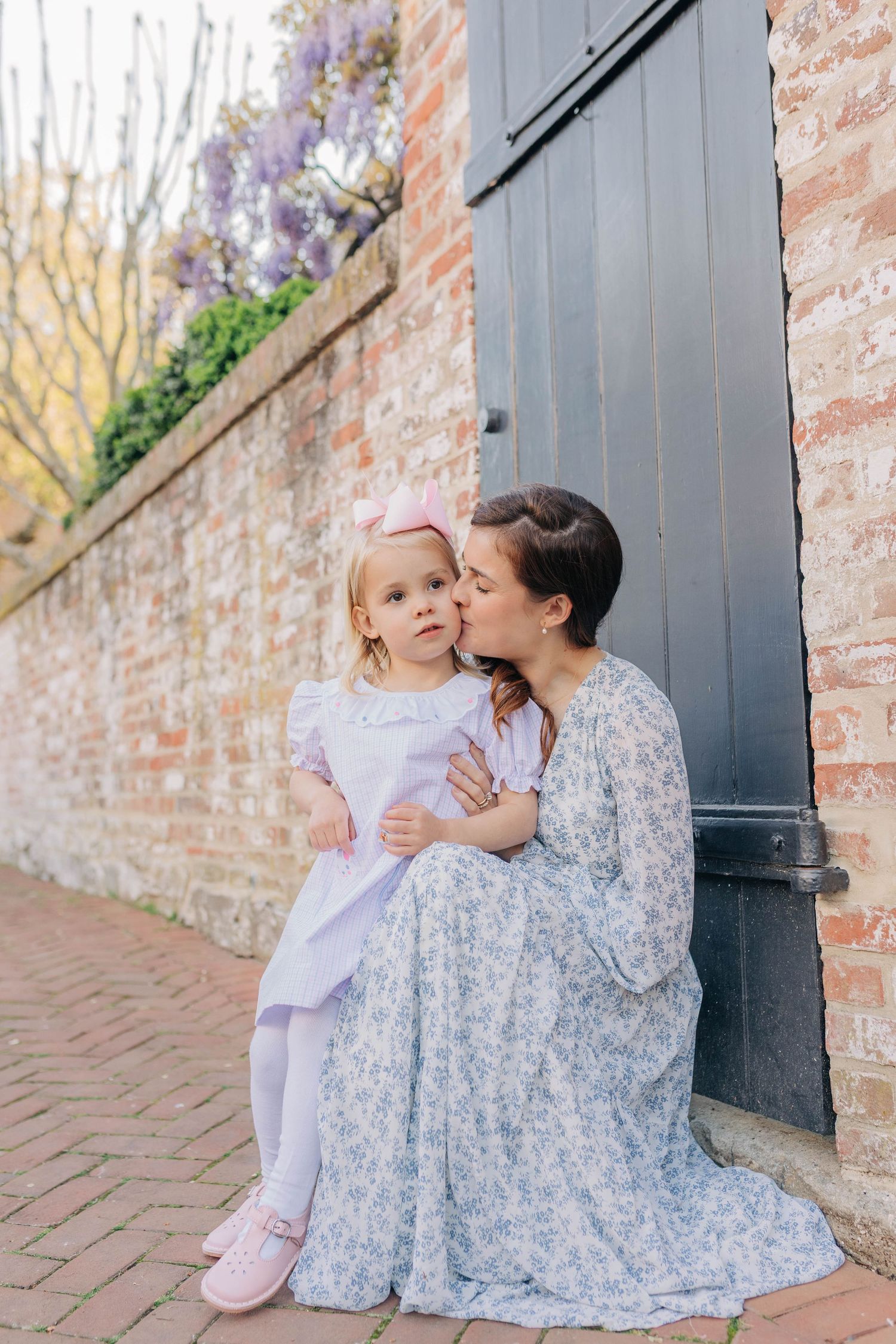 Mother in floral dress kisses toddler girl in pink bow and white dress by brick wall with wisteria.