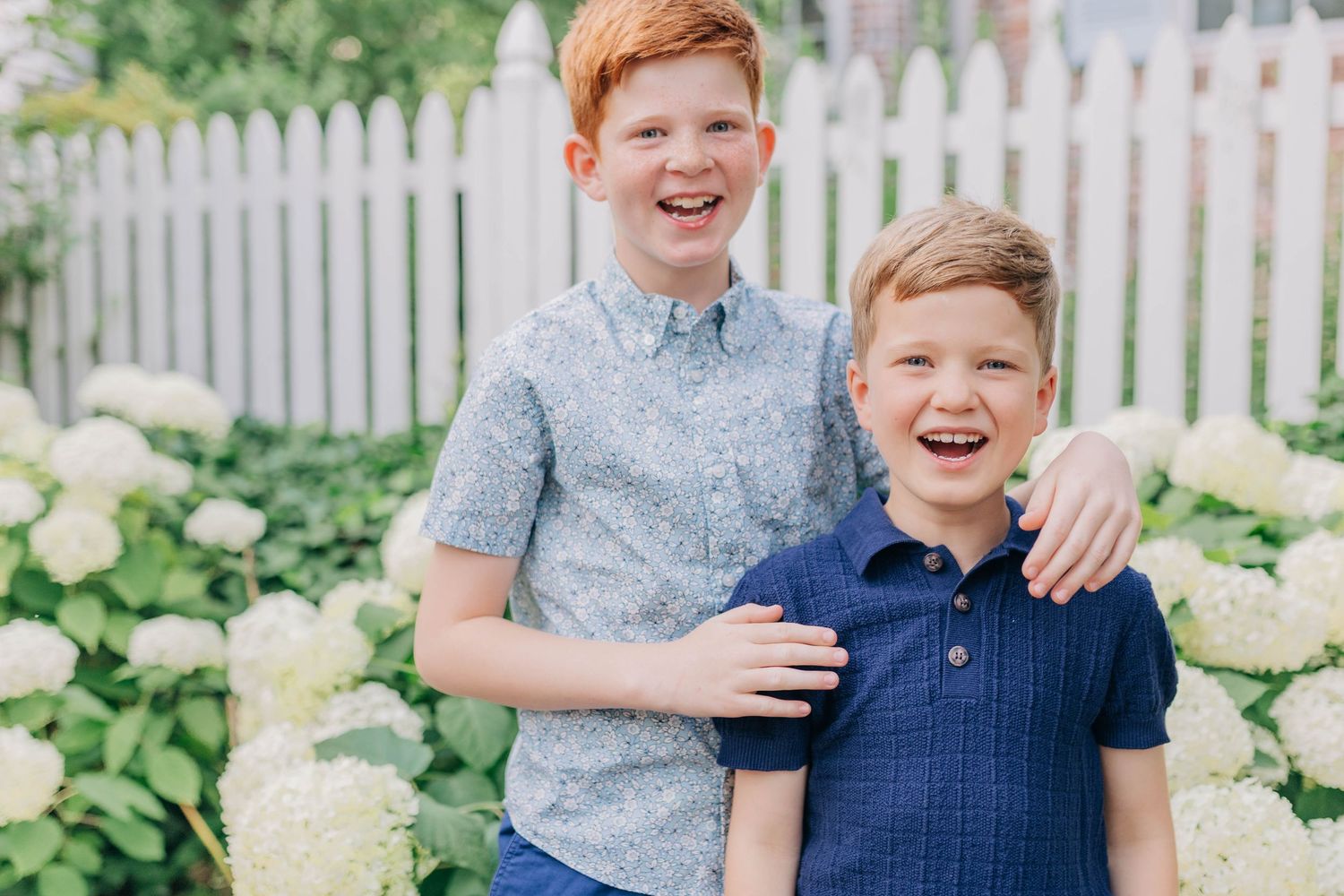 Two smiling red-haired boys pose together in front of white hydrangeas and a picket fence outdoors.