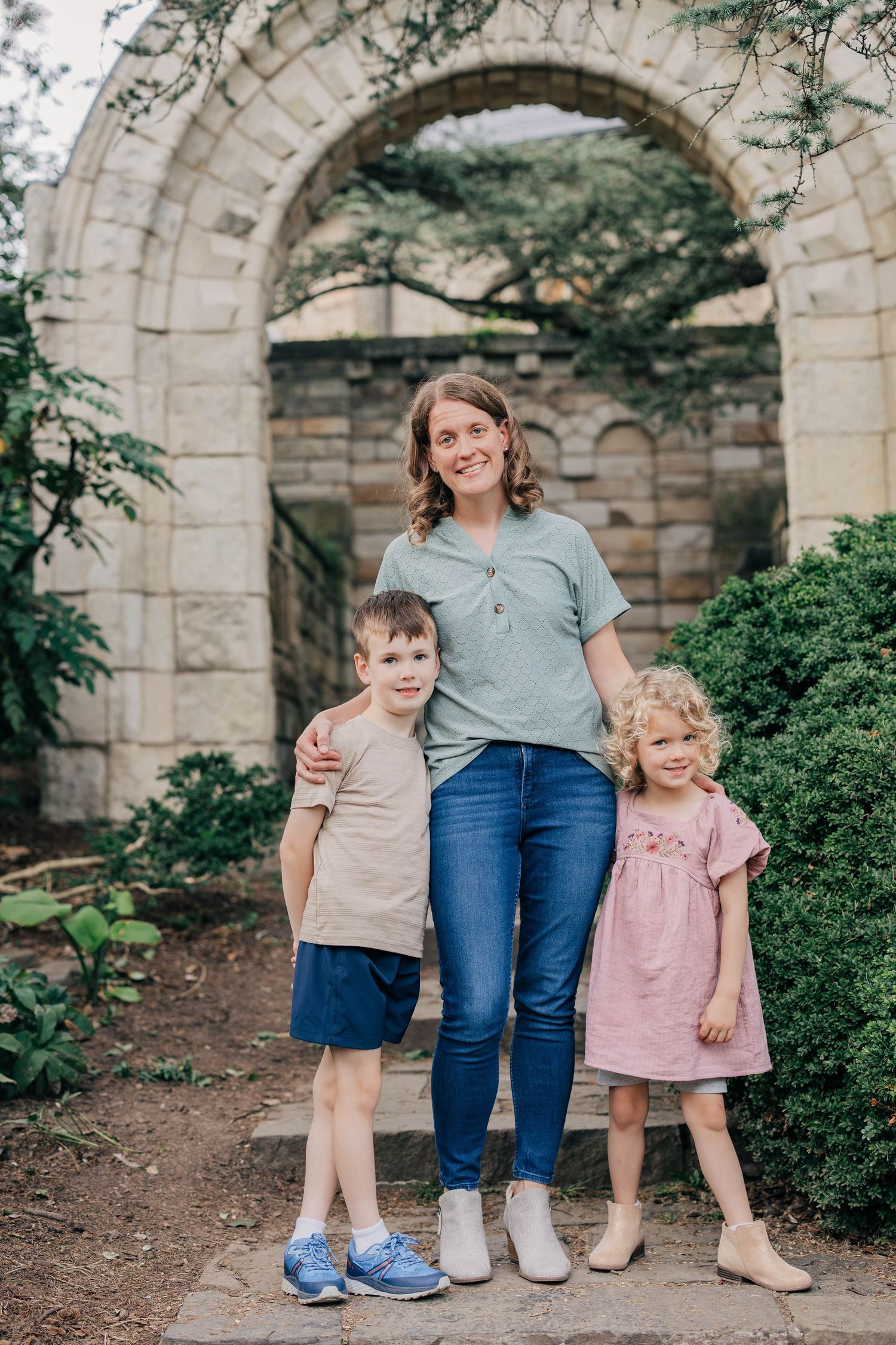 A smiling woman in jeans stands with a young boy and a girl in a pink dress before a stone archway in a garden.