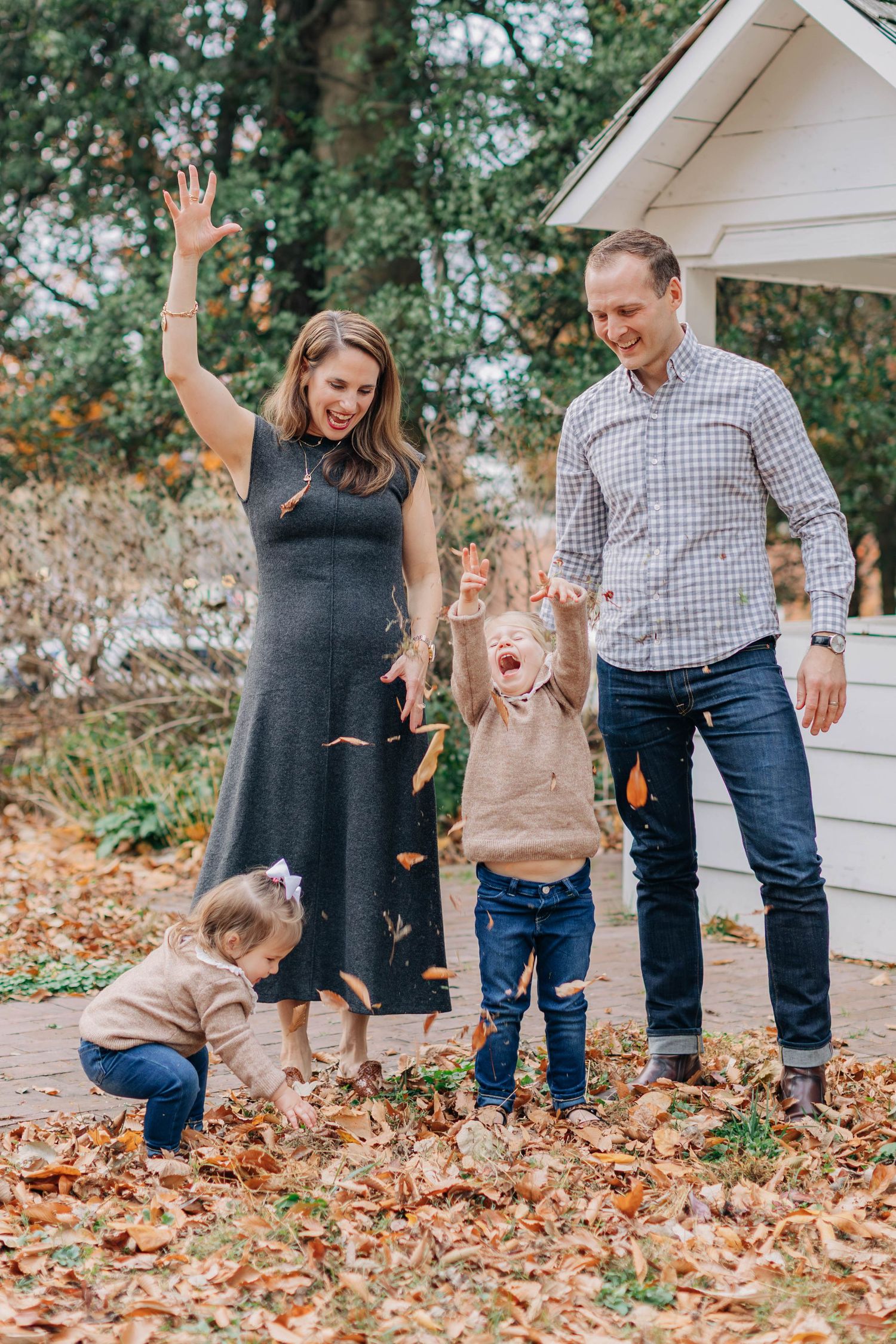 Family of four joyfully tossing autumn leaves outdoors, with pregnant mother, father, toddler, and baby in fall setting.