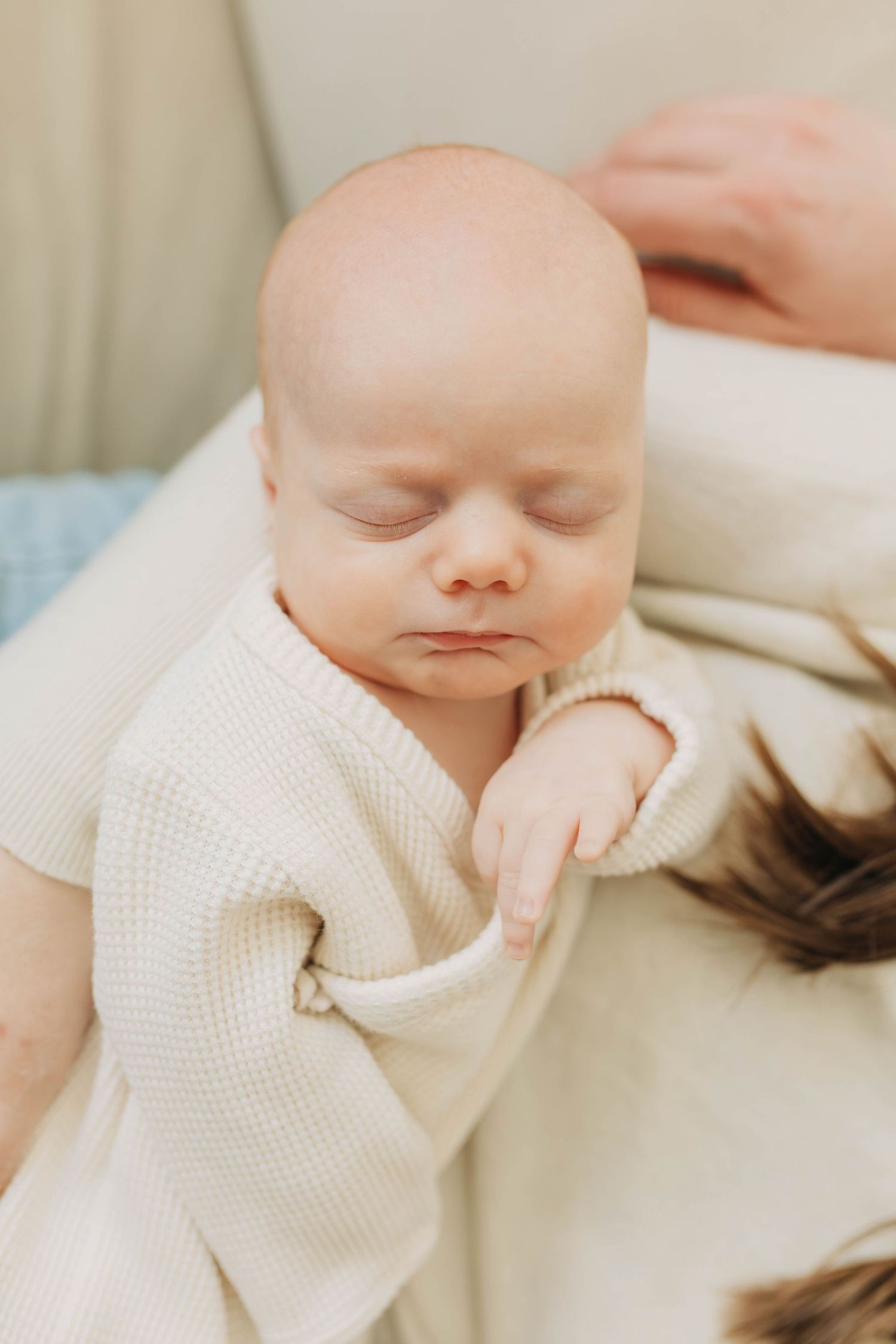 Sleeping newborn baby in cream knit outfit held by mother.
