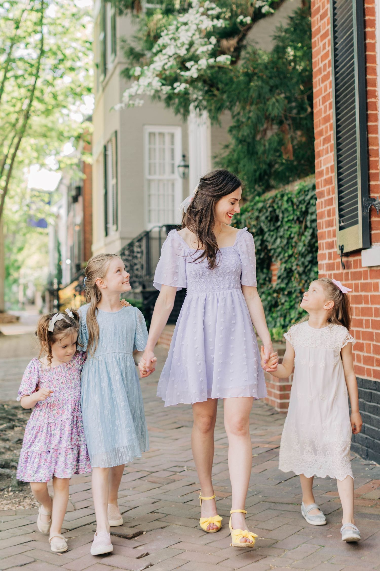 A smiling mother in a lavender dress walks hand-in-hand with three young girls in pastel dresses on a charming brick sidewalk.