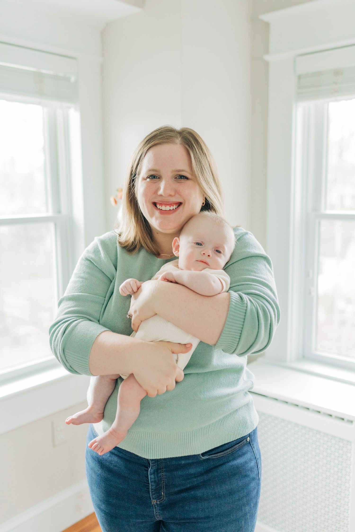 Parent in mint sweater embraces baby in sunny white room.