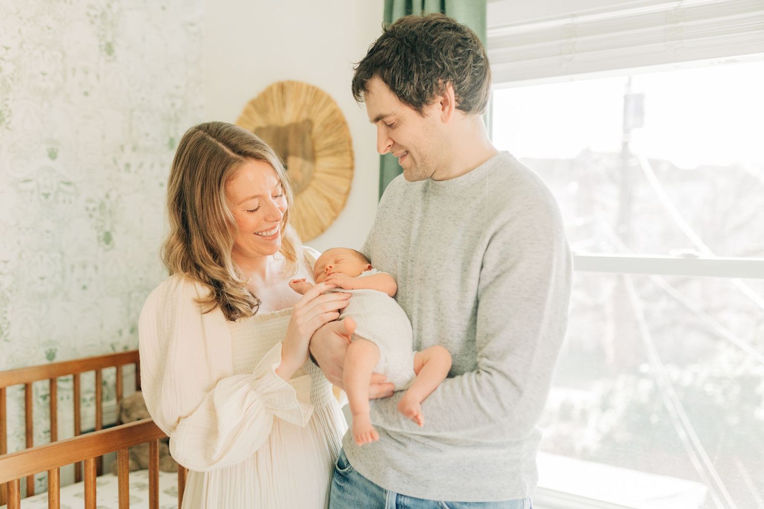 Couple in casual attire smiling while holding their newborn baby near a window.
