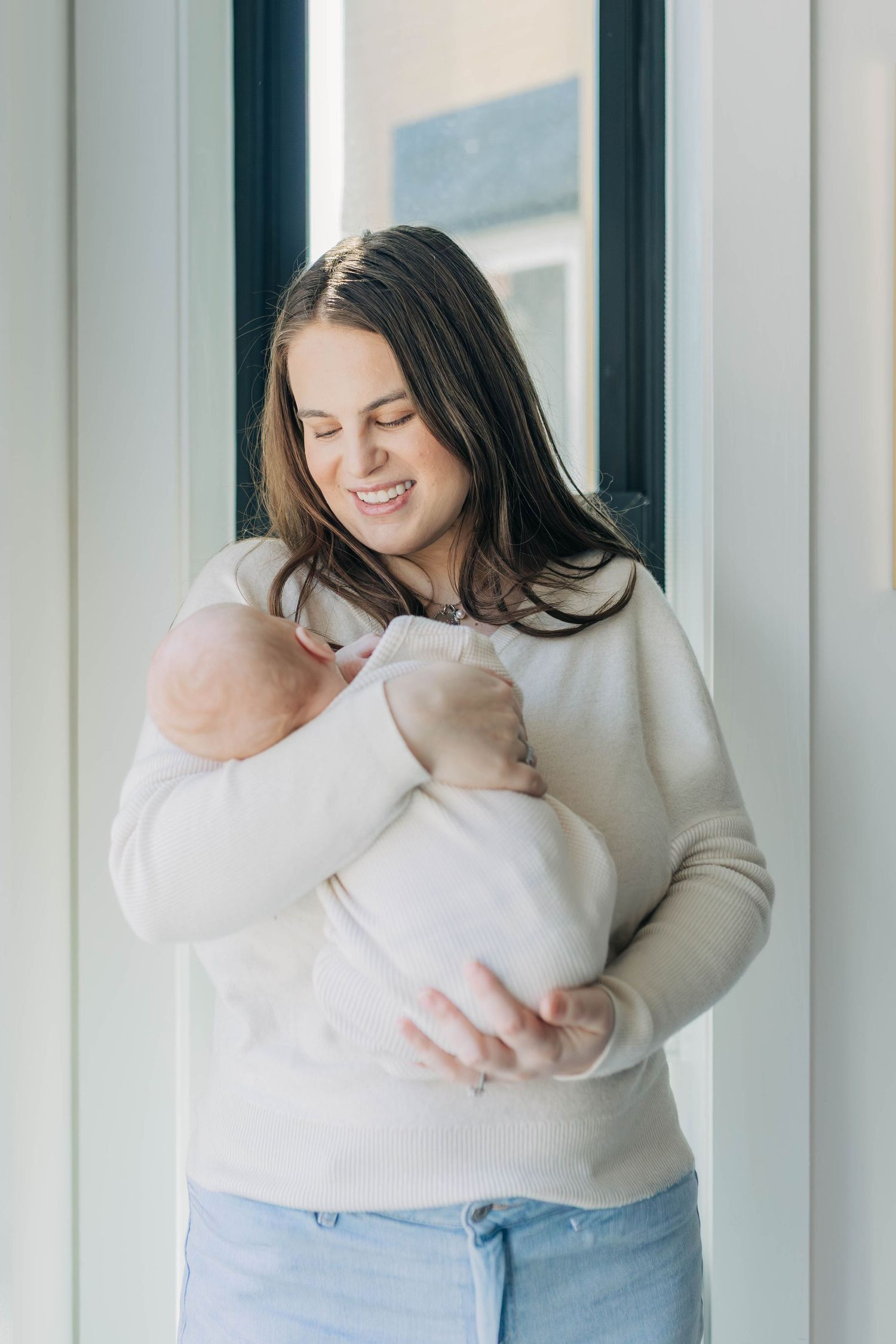 Happy brunette mother in cream sweater and jeans tenderly cradling swaddled newborn baby by window.