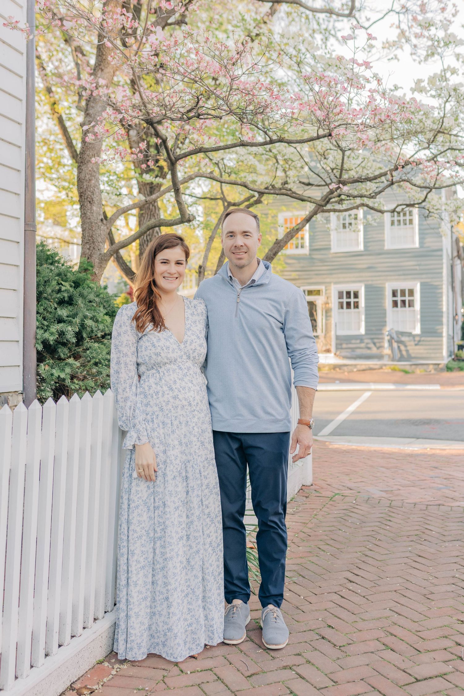 A smiling couple in coordinating blue outfits stands together on a brick sidewalk near blooming spring trees.