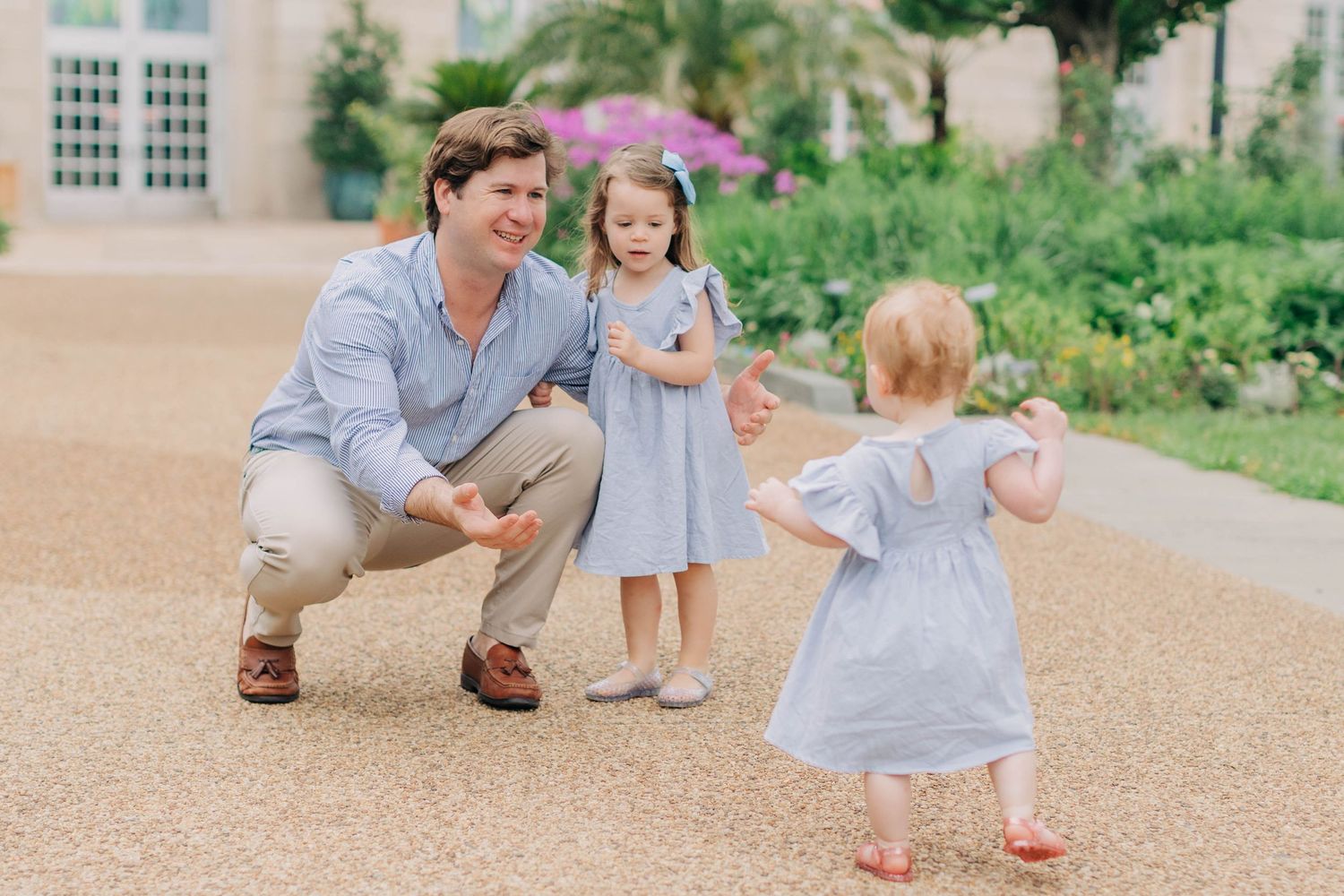 A father crouches down smiling as his two young daughters in blue dresses run toward him outdoors.