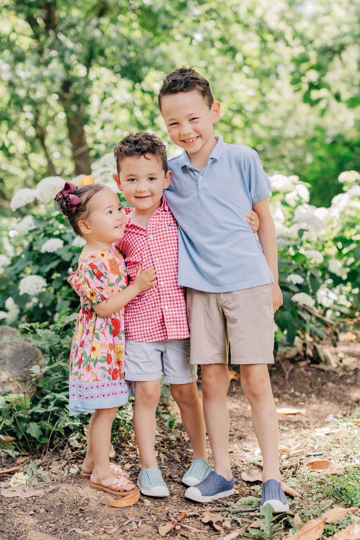 Three young siblings smile together outdoors among white hydrangeas, dressed in colorful summer outfits.