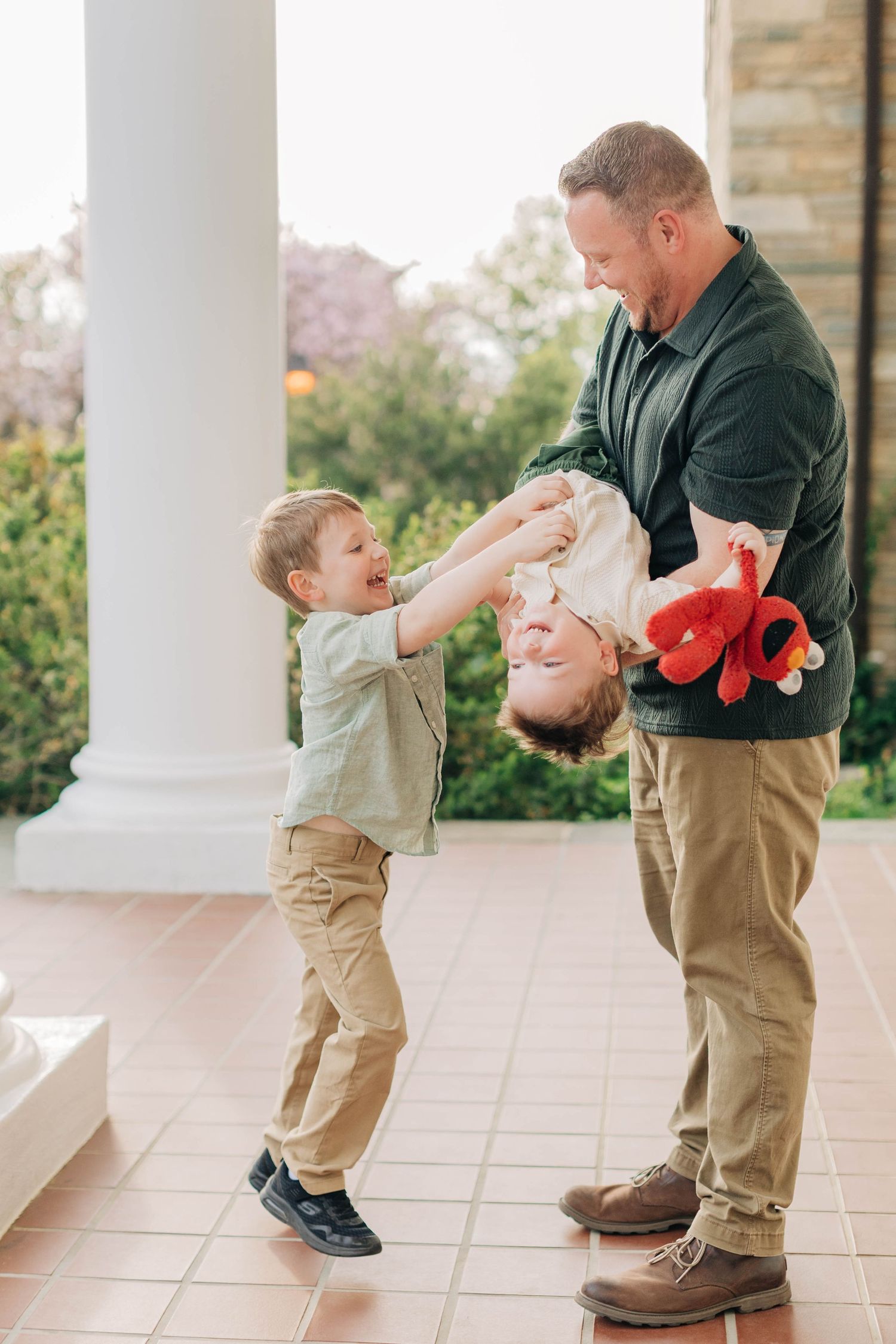 Father playfully holds toddler upside down while older son laughs outdoors.