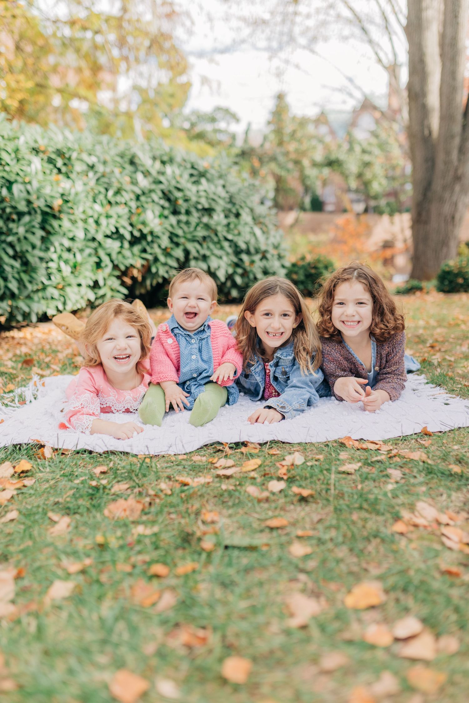 Four happy children lie on a blanket in a fall garden, smiling and laughing among autumn leaves on green grass.