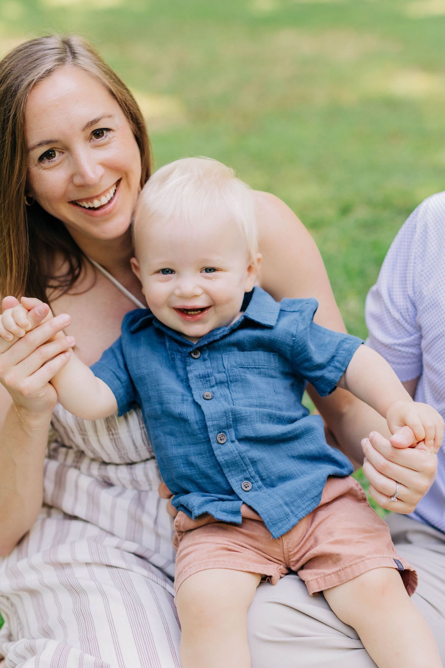 Smiling mother holding happy baby boy in blue shirt and pink shorts outdoors on green grass.