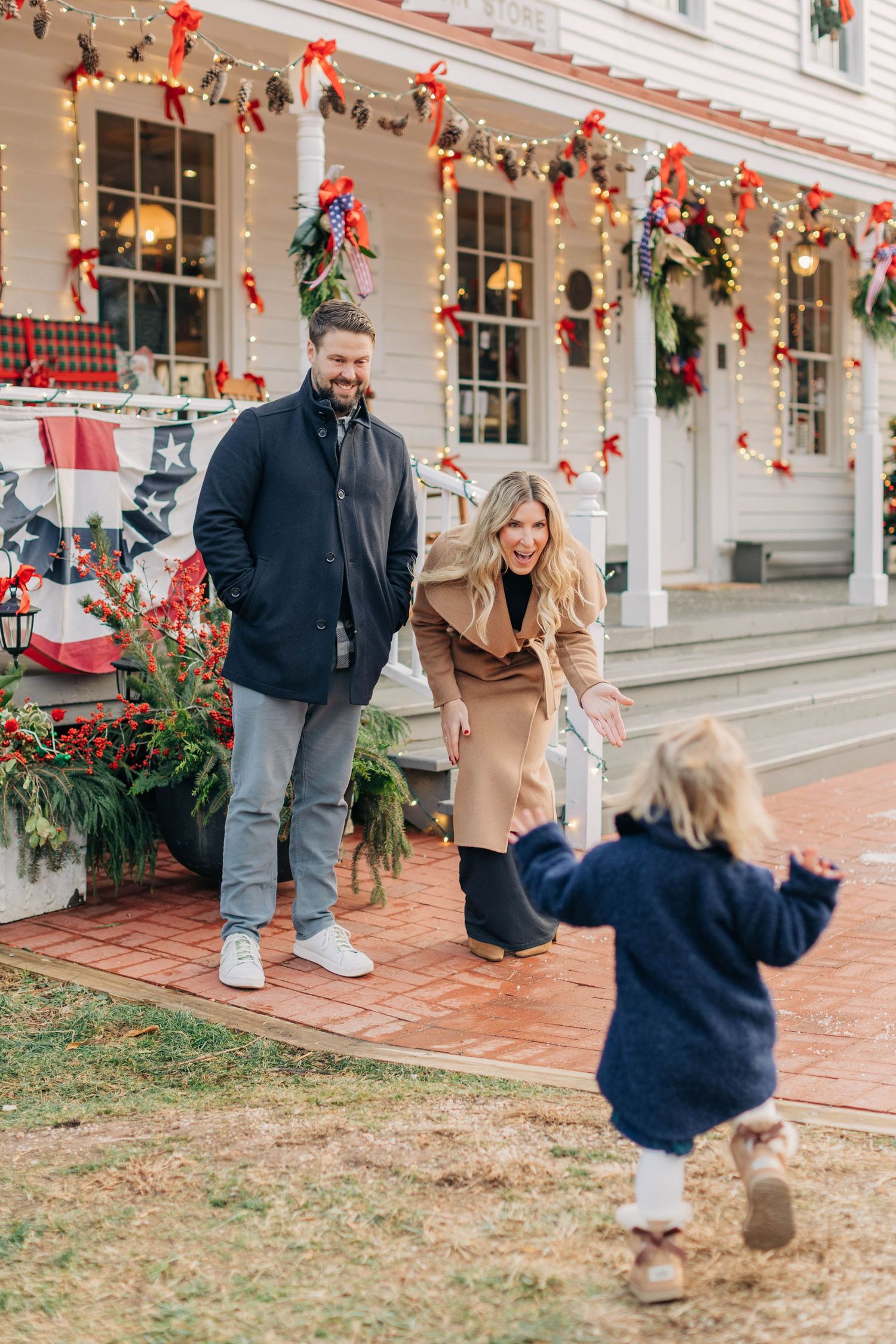Family enjoying Christmas decorations outside a festive home, with a toddler running toward smiling parents.