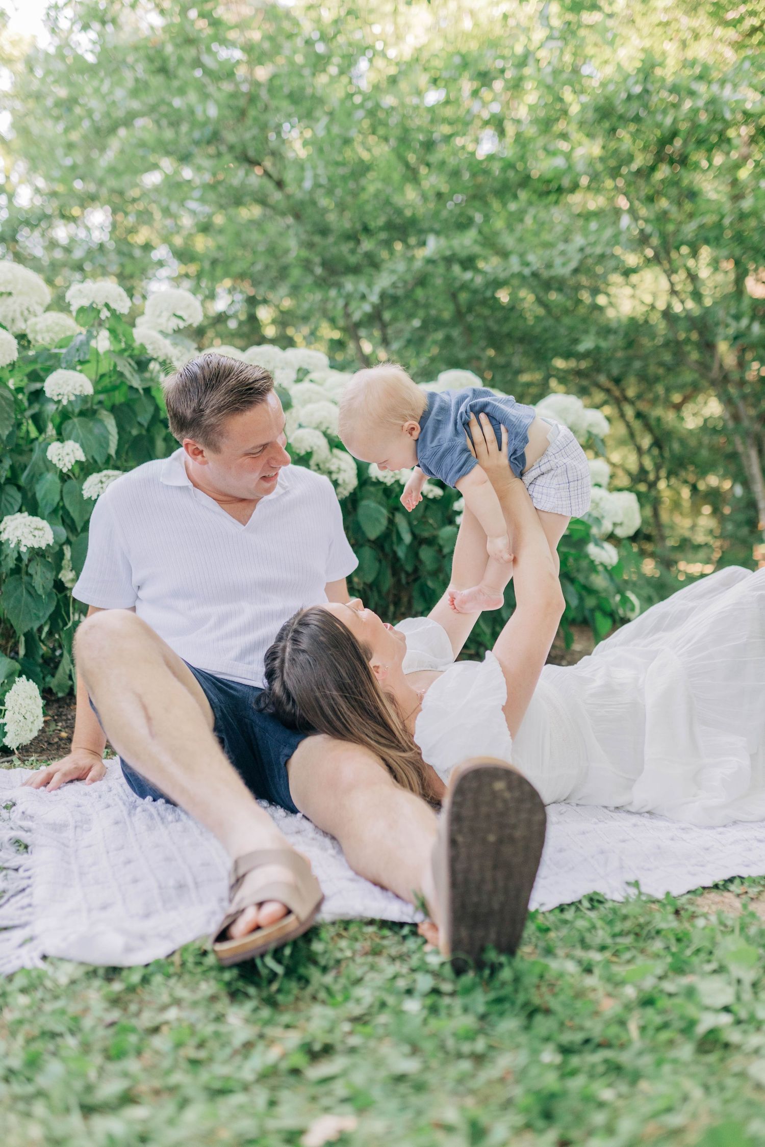 Family shares playful moment on white blanket surrounded by flowering bushes in garden.