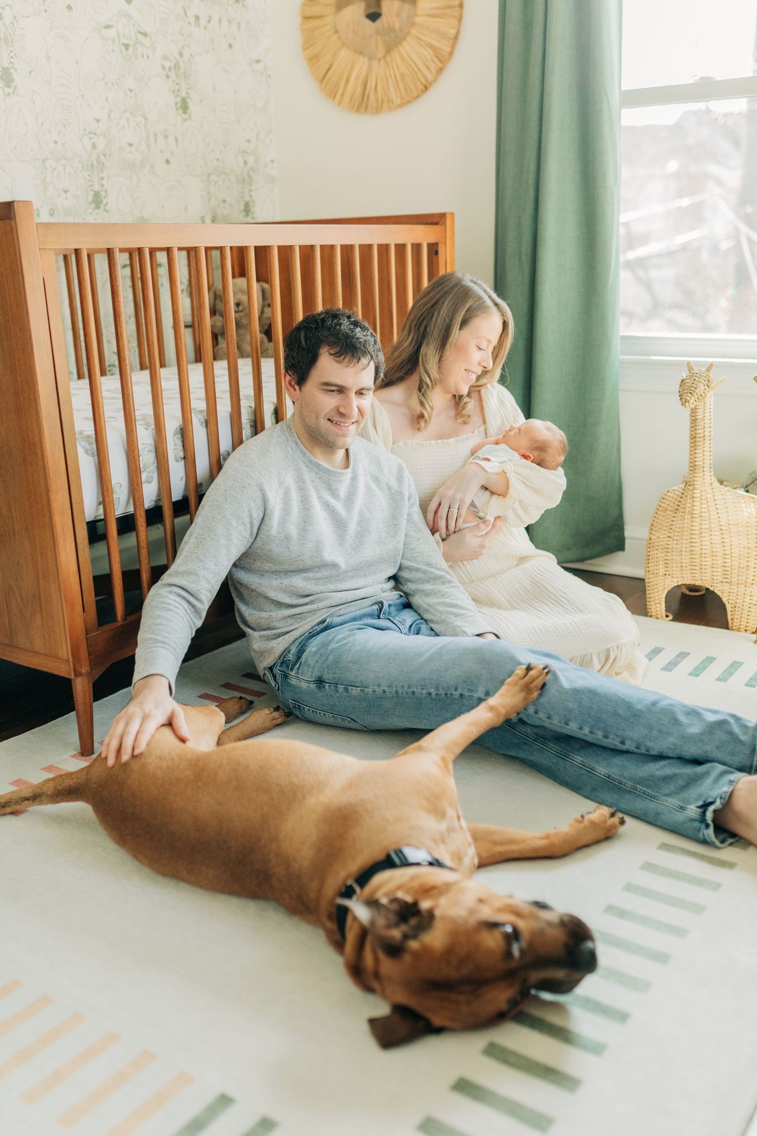 Parents sit on striped rug with sleeping dog while holding newborn in nursery.