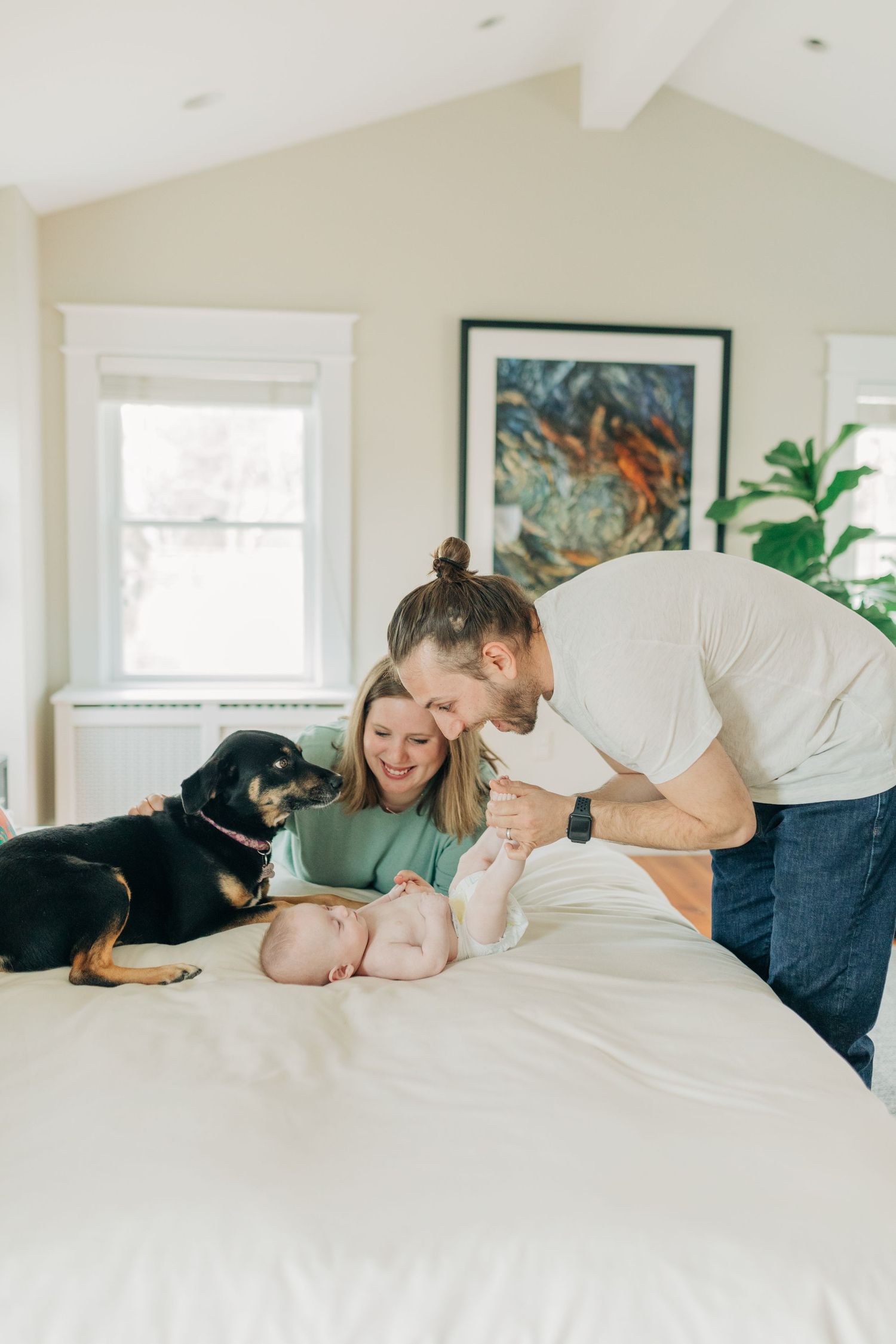 Family and black dog sharing sweet moments with baby on white bed in sunlit room.