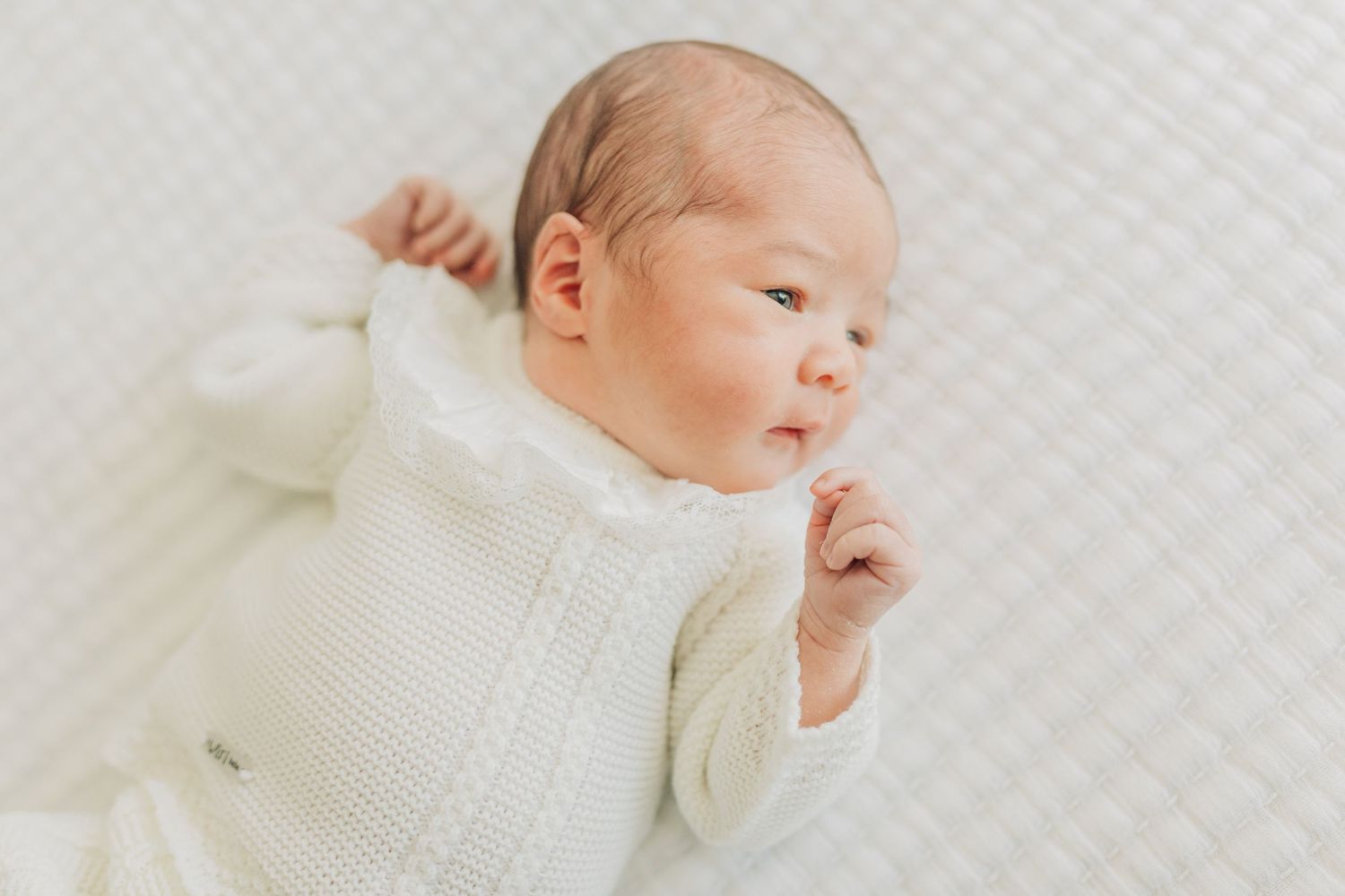 A newborn baby wearing a white knit sweater rests peacefully on white bedding.